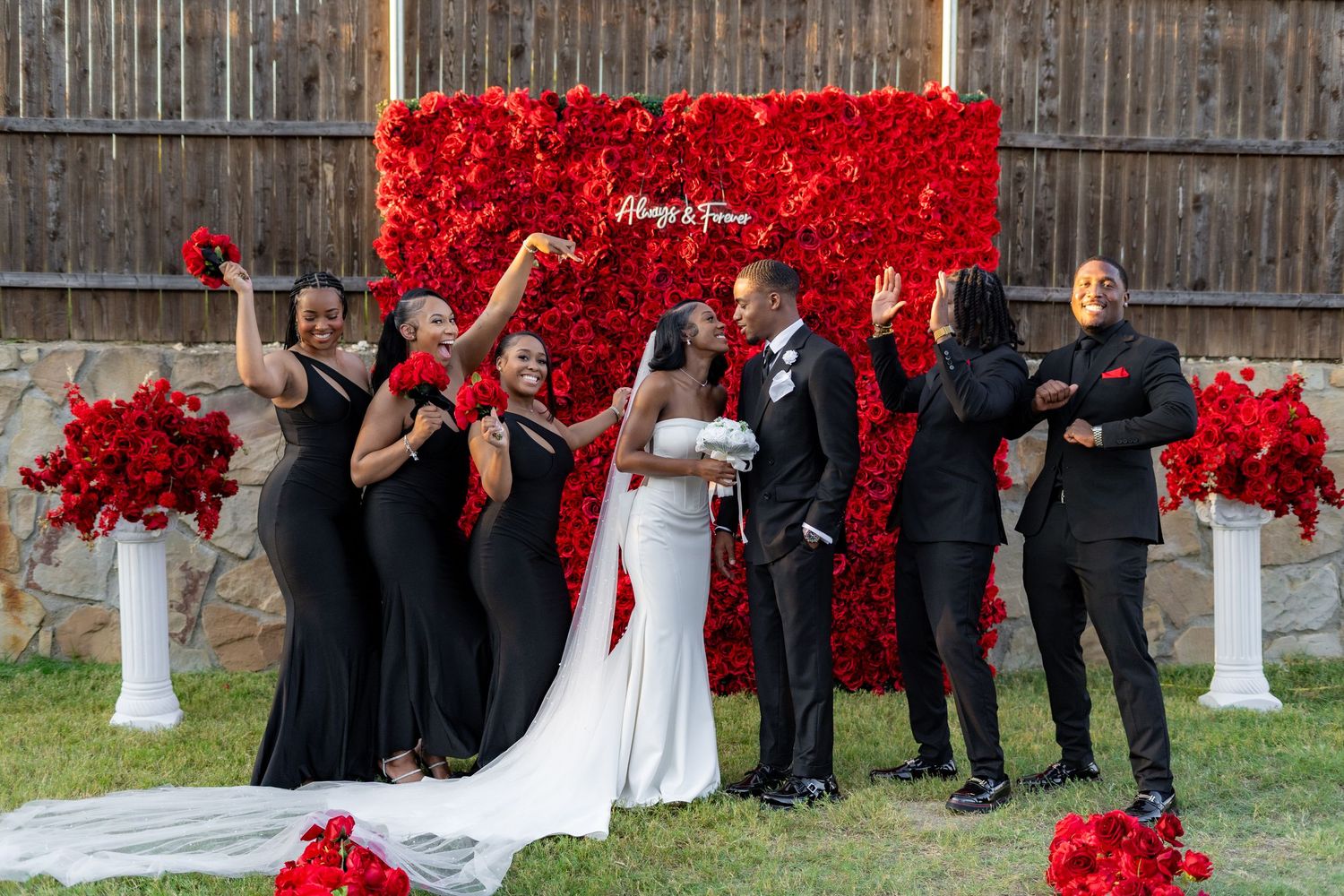 Wedding party celebrates in front of a red rose flower wall with Always & Forever neon sign outdoors.