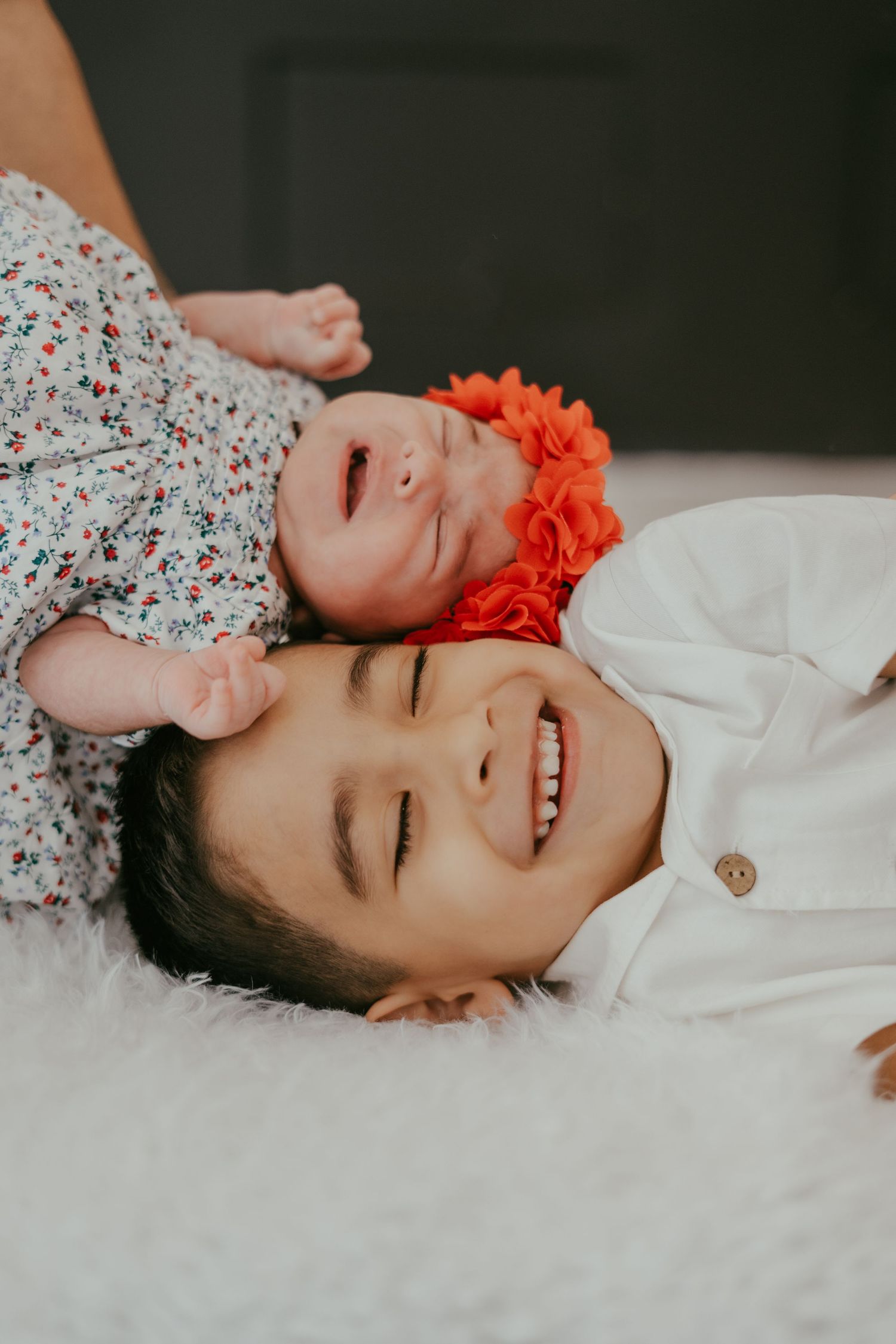 Laughing older child lies on white fur blanket as newborn baby girl with orange flower headband rests on their chest.