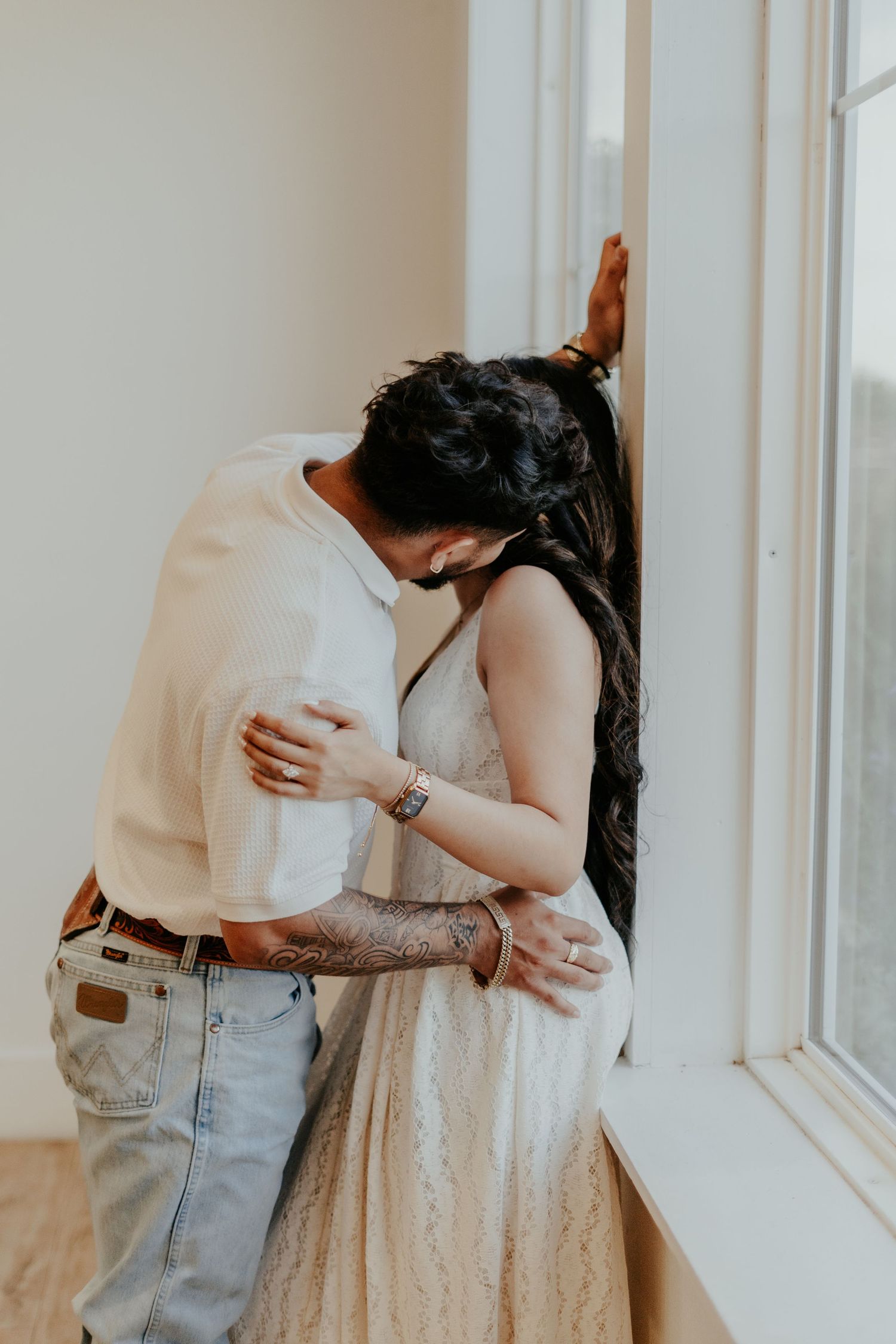 Couple sharing an intimate moment by a window, man with tattoos embracing woman in a lace dress.