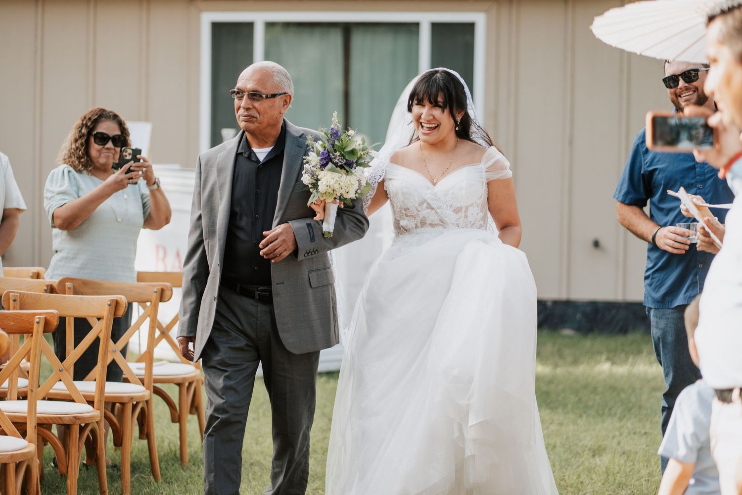 Bride in white lace gown walks down outdoor aisle with father during sunny wedding ceremony.