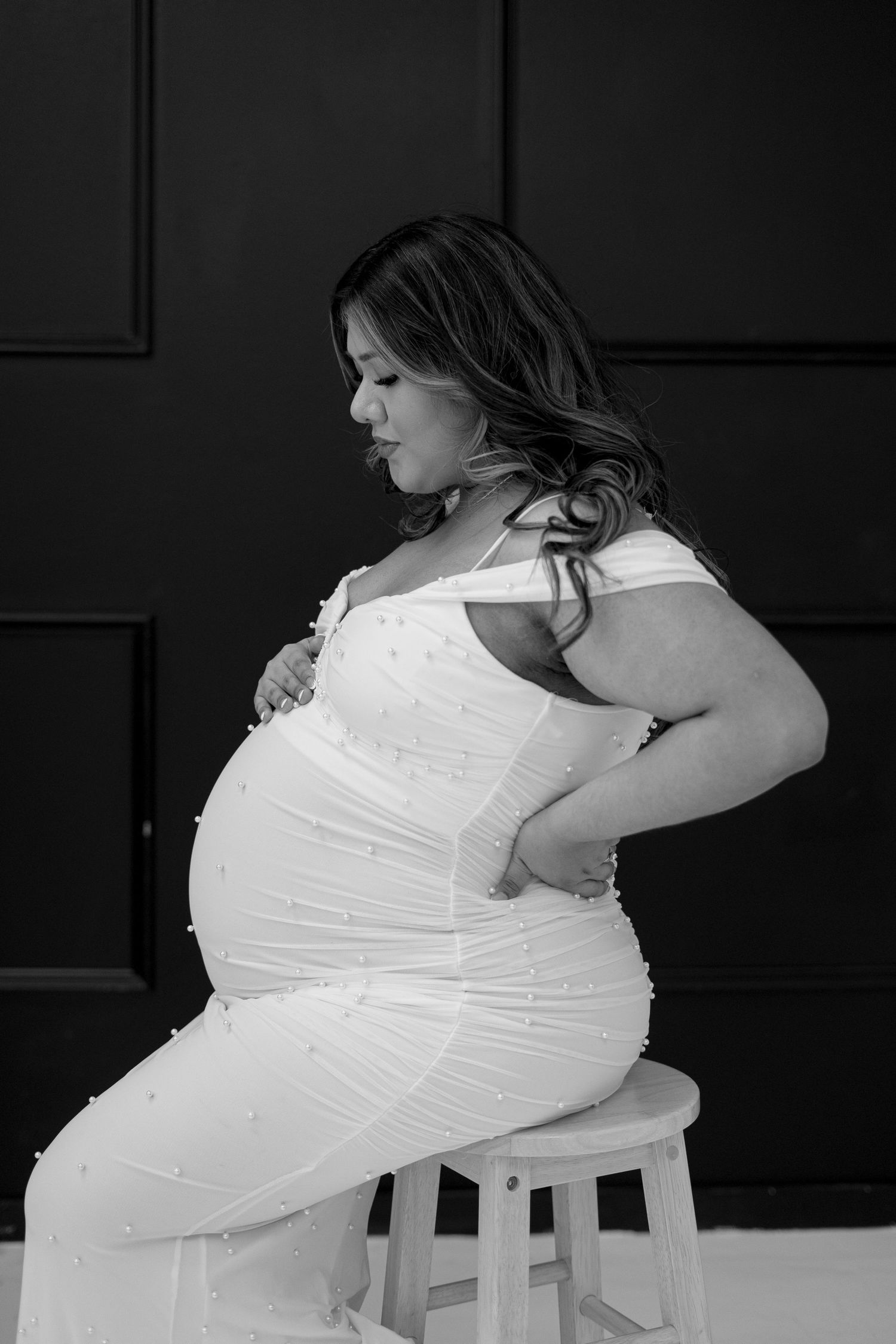 Black and white portrait of pregnant woman in white pearl gown sitting on stool in studio.