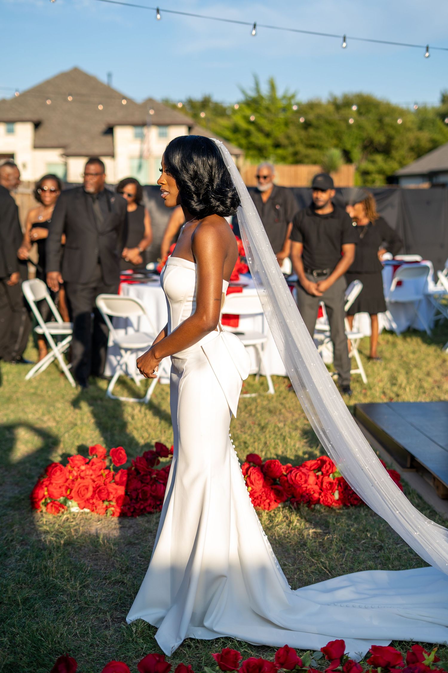 Elegant bride in fitted white gown with cathedral veil stands among red roses at outdoor ceremony.