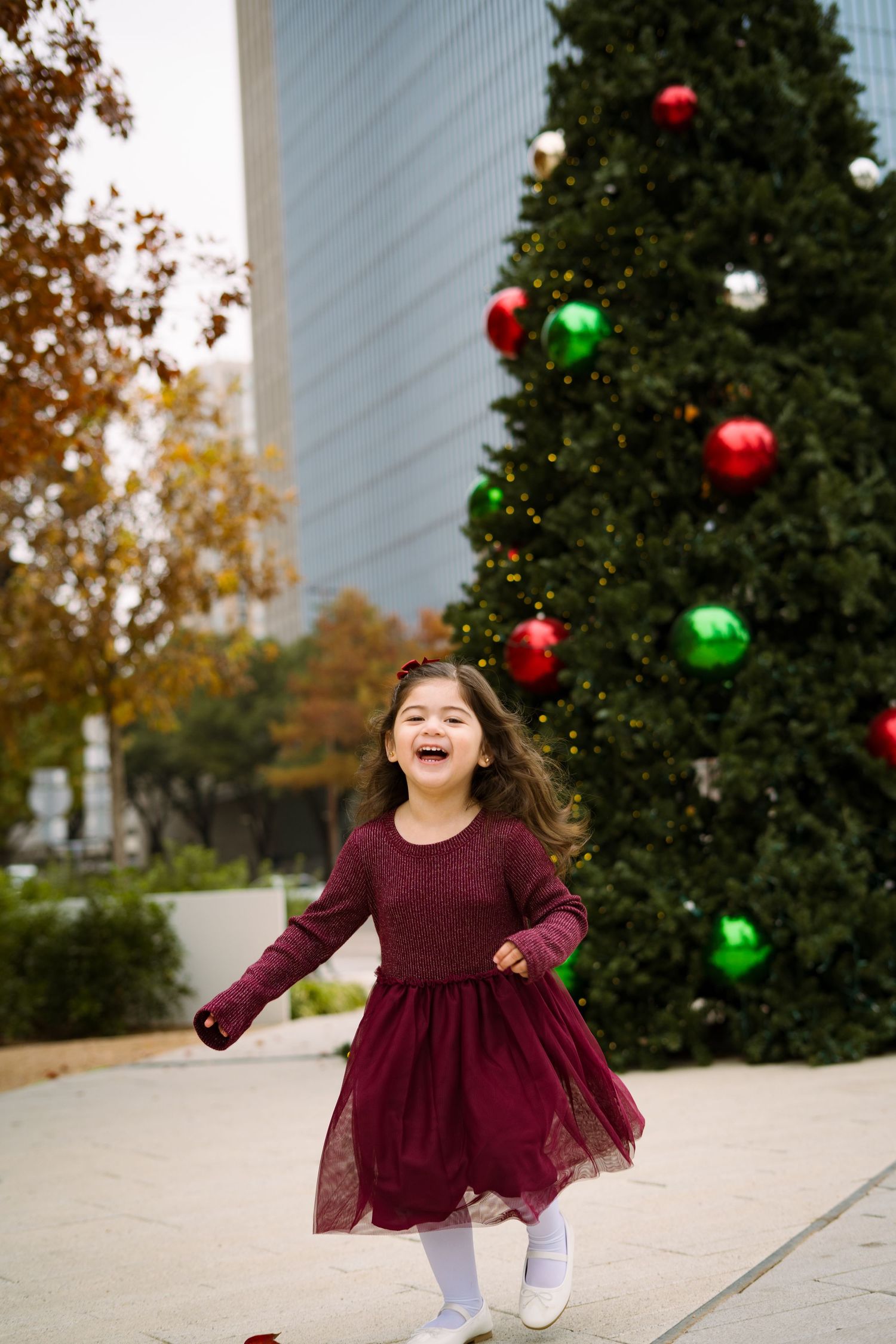 Happy little girl in burgundy dress laughing and running in front of a large outdoor Christmas tree.