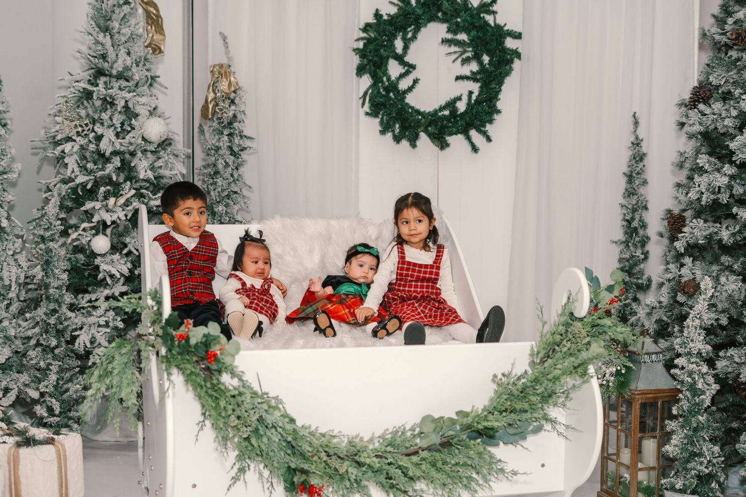 Four young children in holiday outfits pose together in a white sleigh surrounded by Christmas trees.