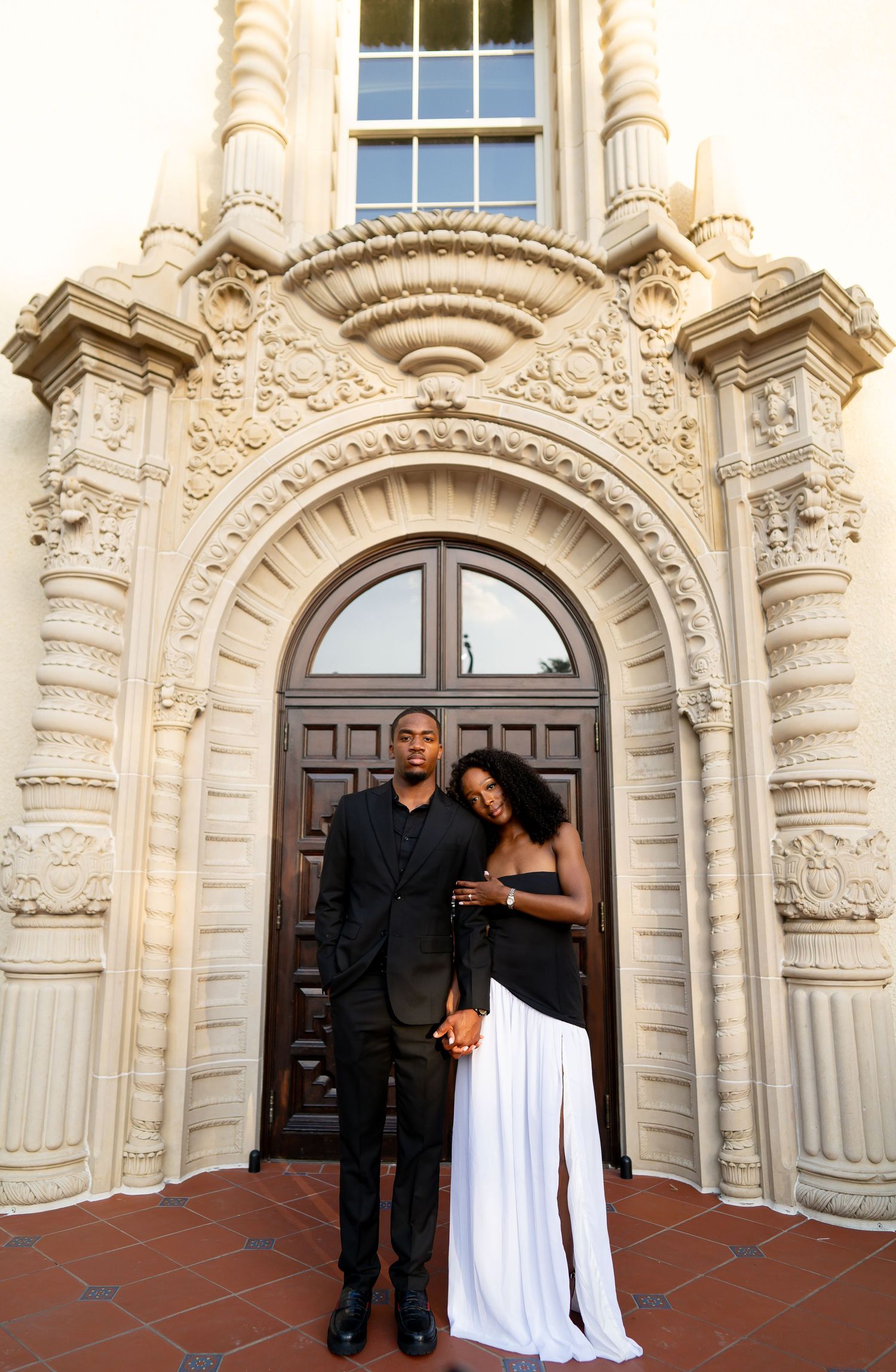 Black couple posing elegantly before ornate arched doorway in coordinated formal black and white attire.
