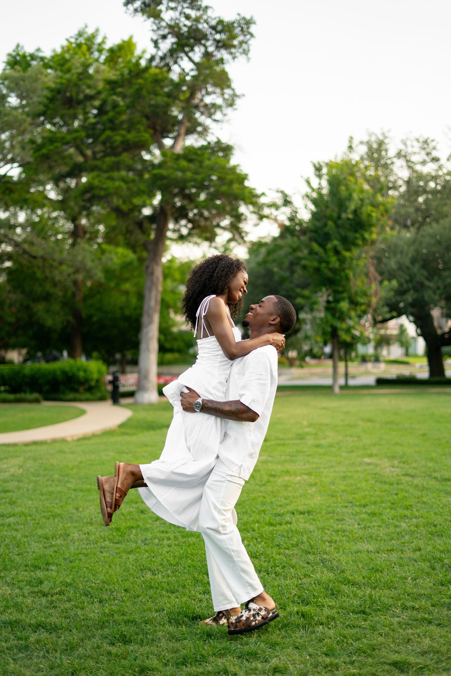 A happy Black man lifts his smiling partner in a white dress as they laugh together in a green park.