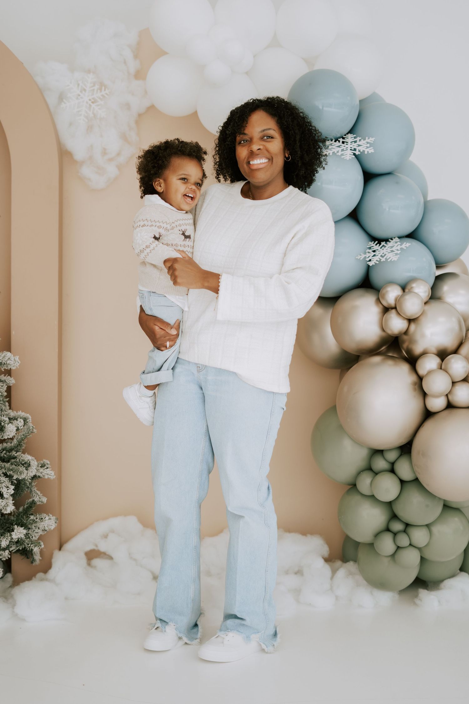 Joyful mother holds toddler in front of blue, gold, and white winter balloon garland display.