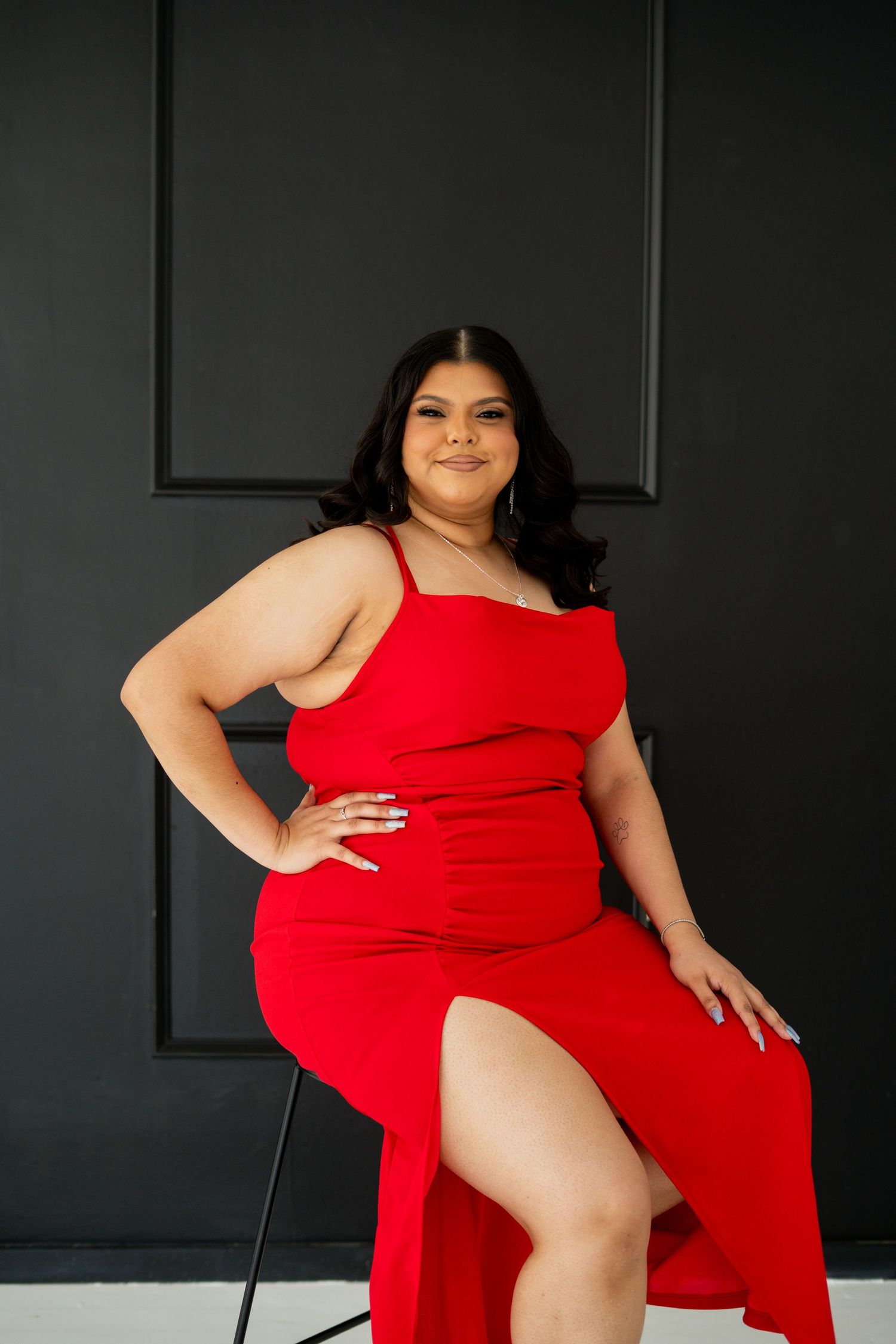 A woman in a red dress sits confidently on a chair against a dark paneled wall.