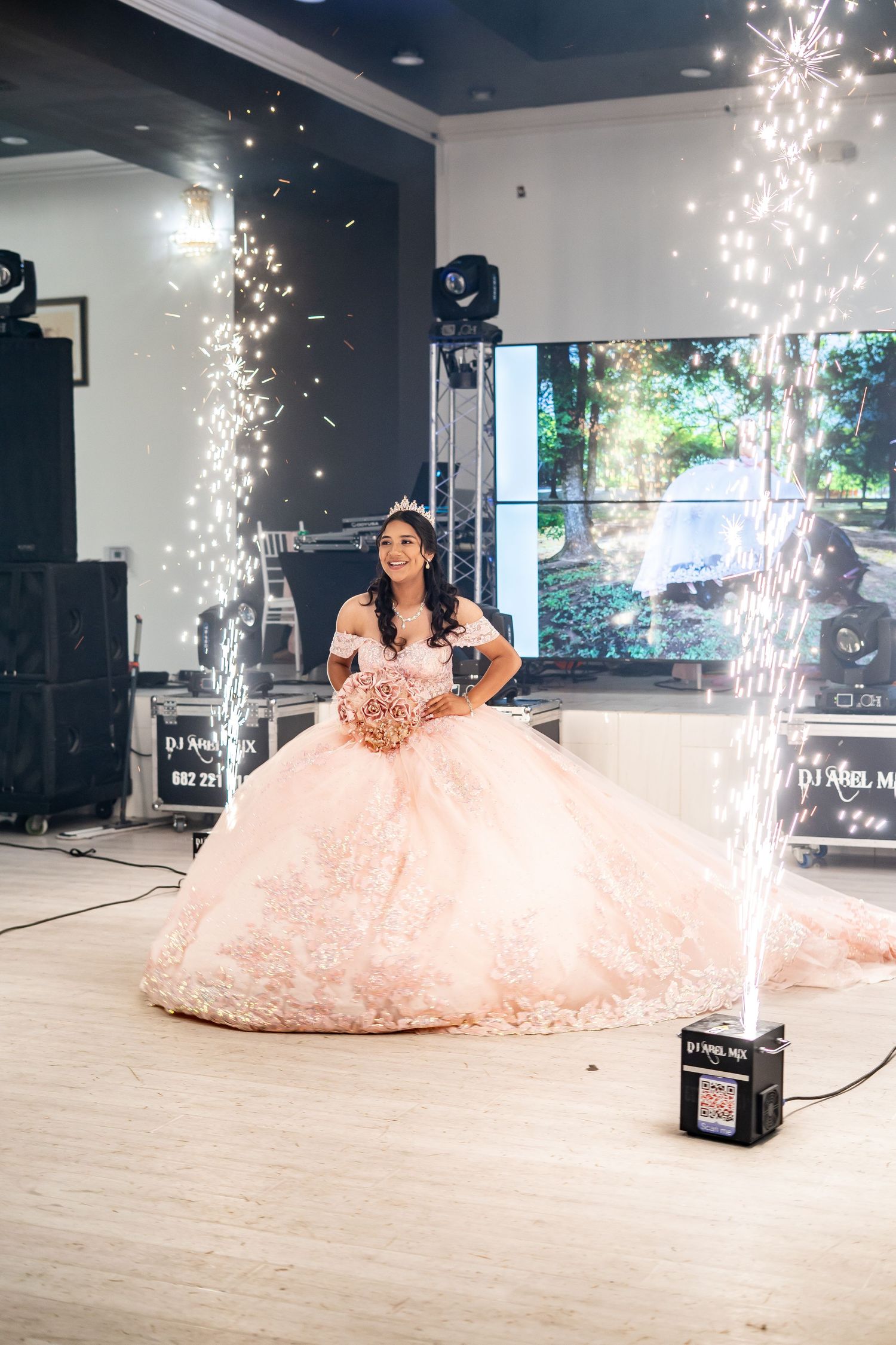 Quinceañera girl in pink ball gown holds bouquet surrounded by sparkling pyrotechnic fountains on dance floor.