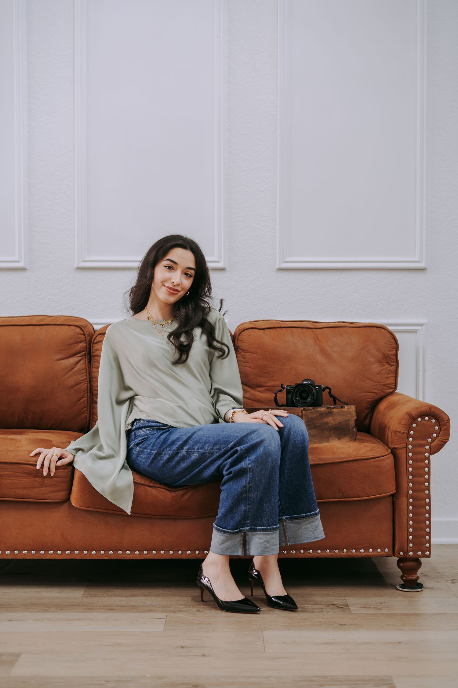 Young woman in green top and jeans sitting on brown leather sofa with camera beside her.