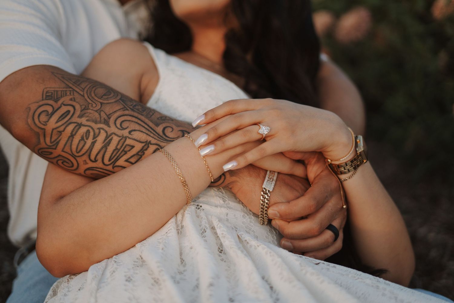 Couple holding hands showing tattoos, bracelets, and an engagement ring, wearing a white dress outdoors.