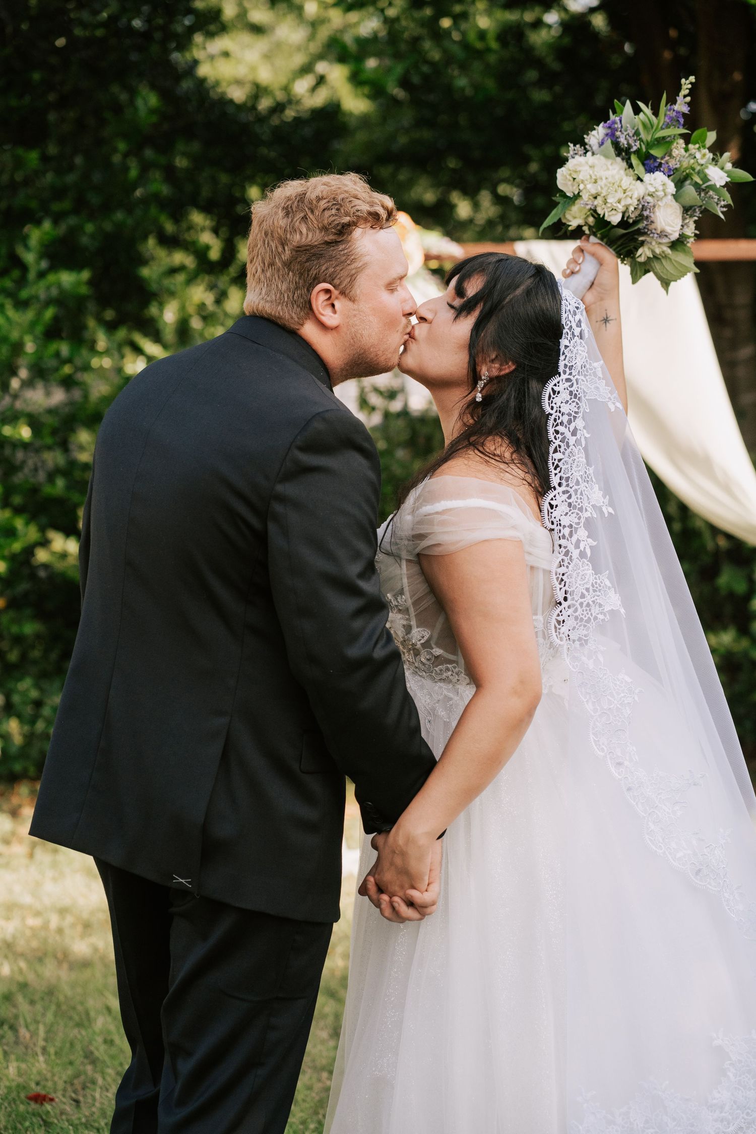 Bride and groom sharing first kiss at outdoor wedding ceremony with bouquet raised.