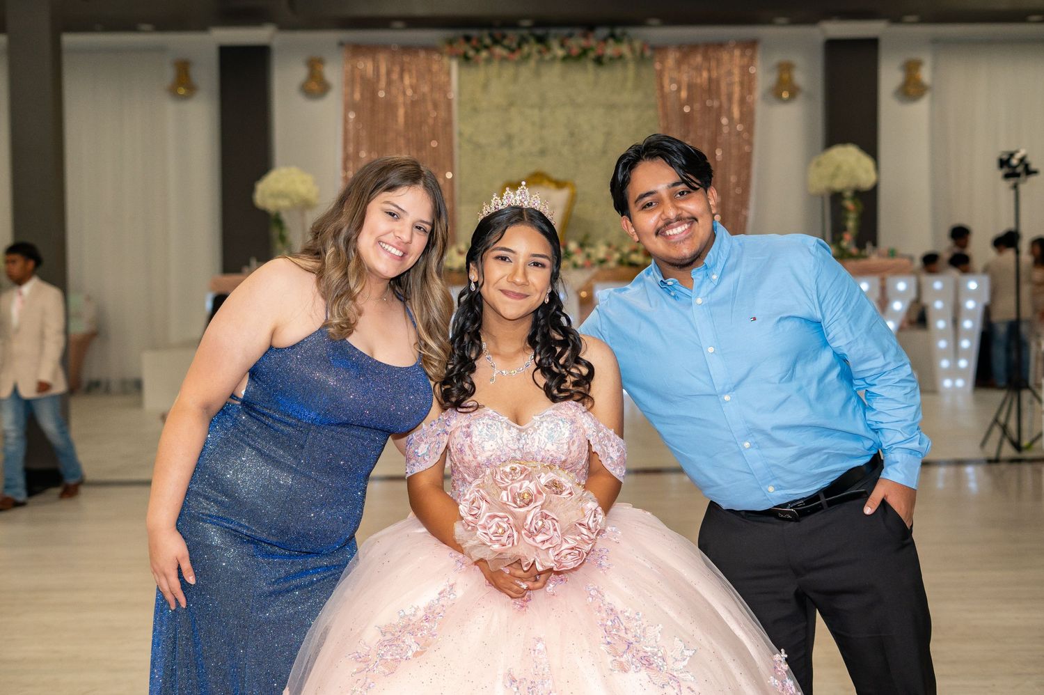 Quinceañera girl in pink gown holds bouquet while posing with two smiling guests at her celebration.