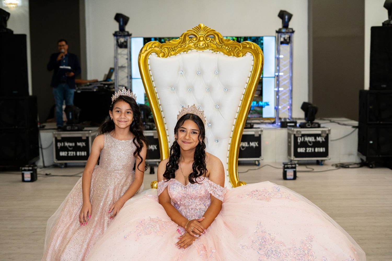 A quinceañera and a young girl pose together in front of a gold throne chair.