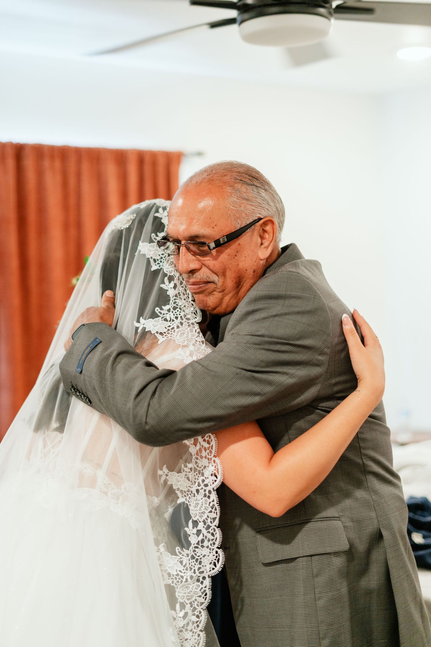 Elderly man in gray suit emotionally hugging a bride in a lace wedding veil during a wedding day moment.