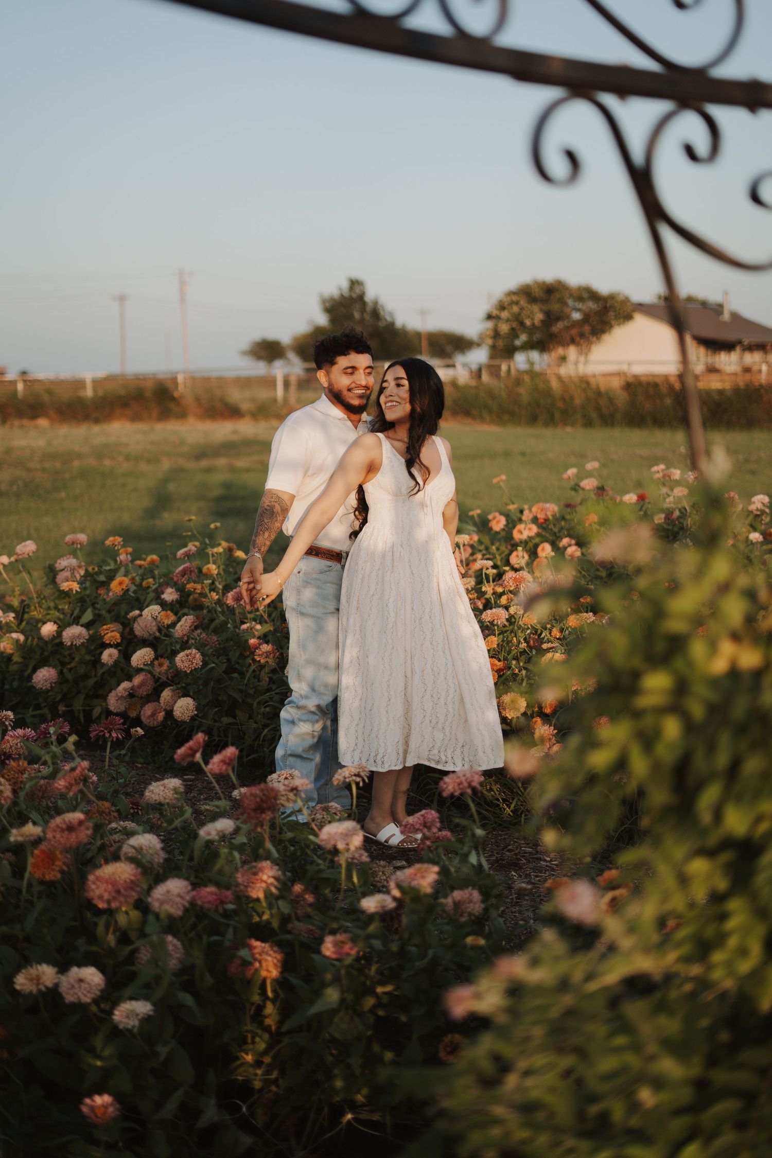 Couple standing romantically among pink dahlia flowers in a garden at golden hour, woman in white dress.