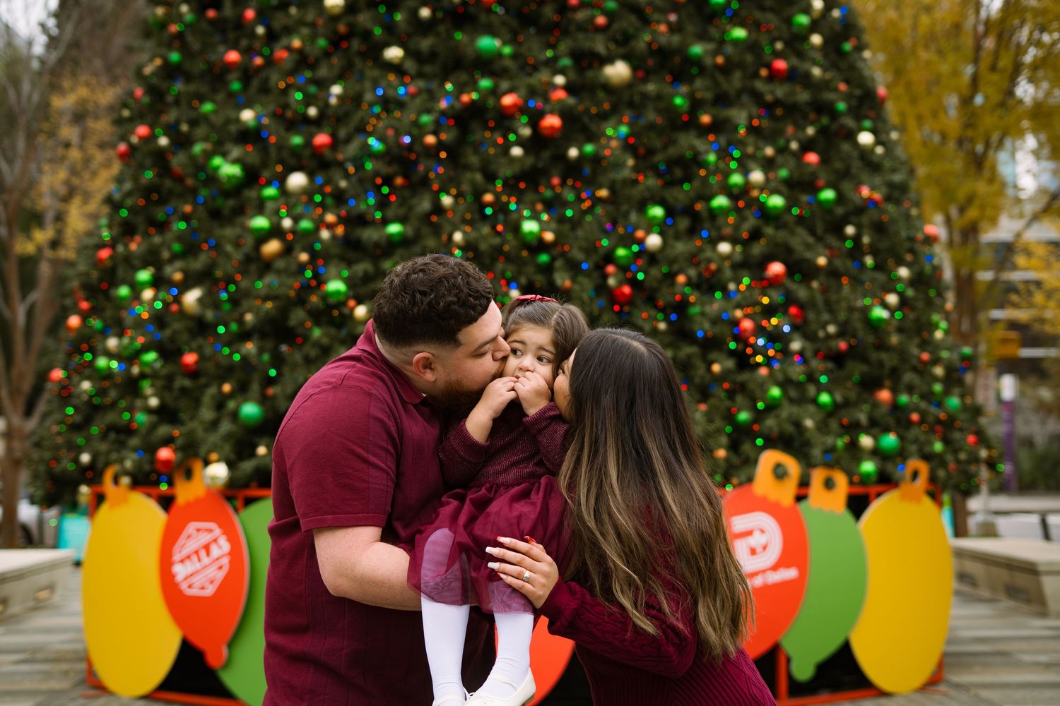 A couple in burgundy sharing a tender kiss with their young child in front of a festive Christmas tree.
