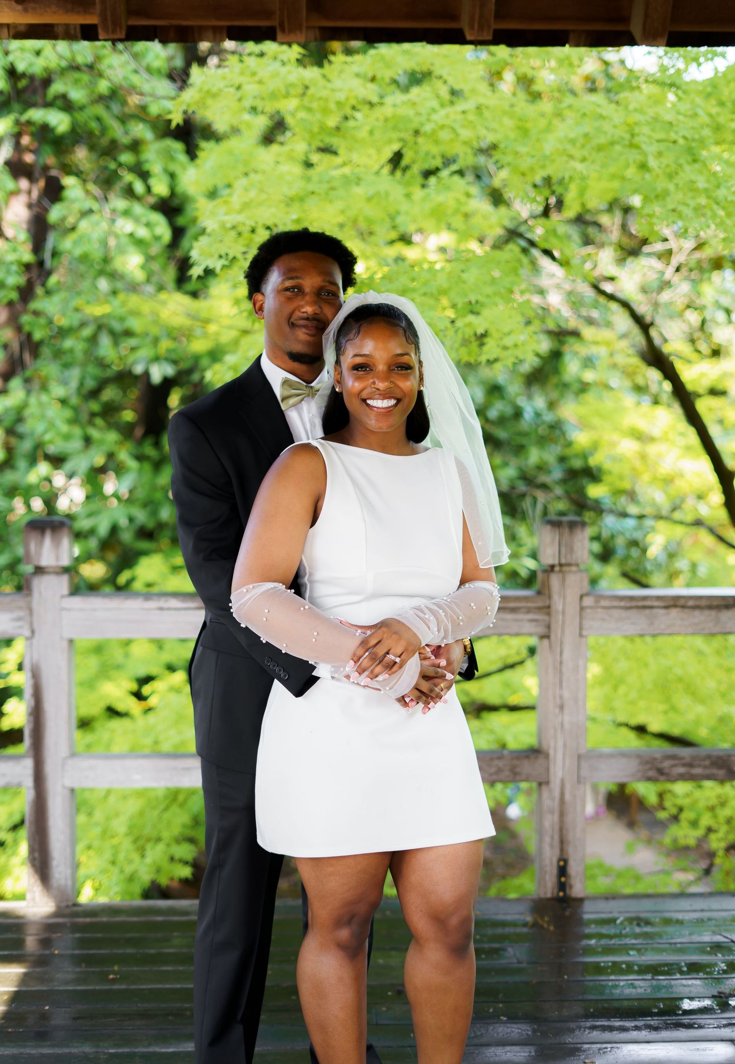 Newlywed couple poses together under wooden pavilion, groom in black tuxedo embracing bride in short white dress.