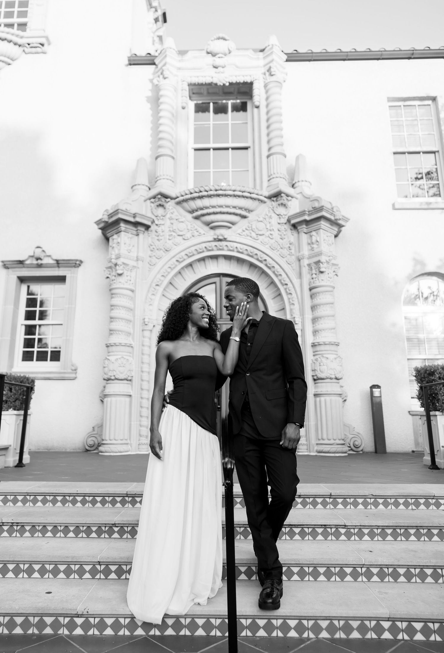 Black and white photo of a couple sharing a whisper on decorative steps before a grand historic building.