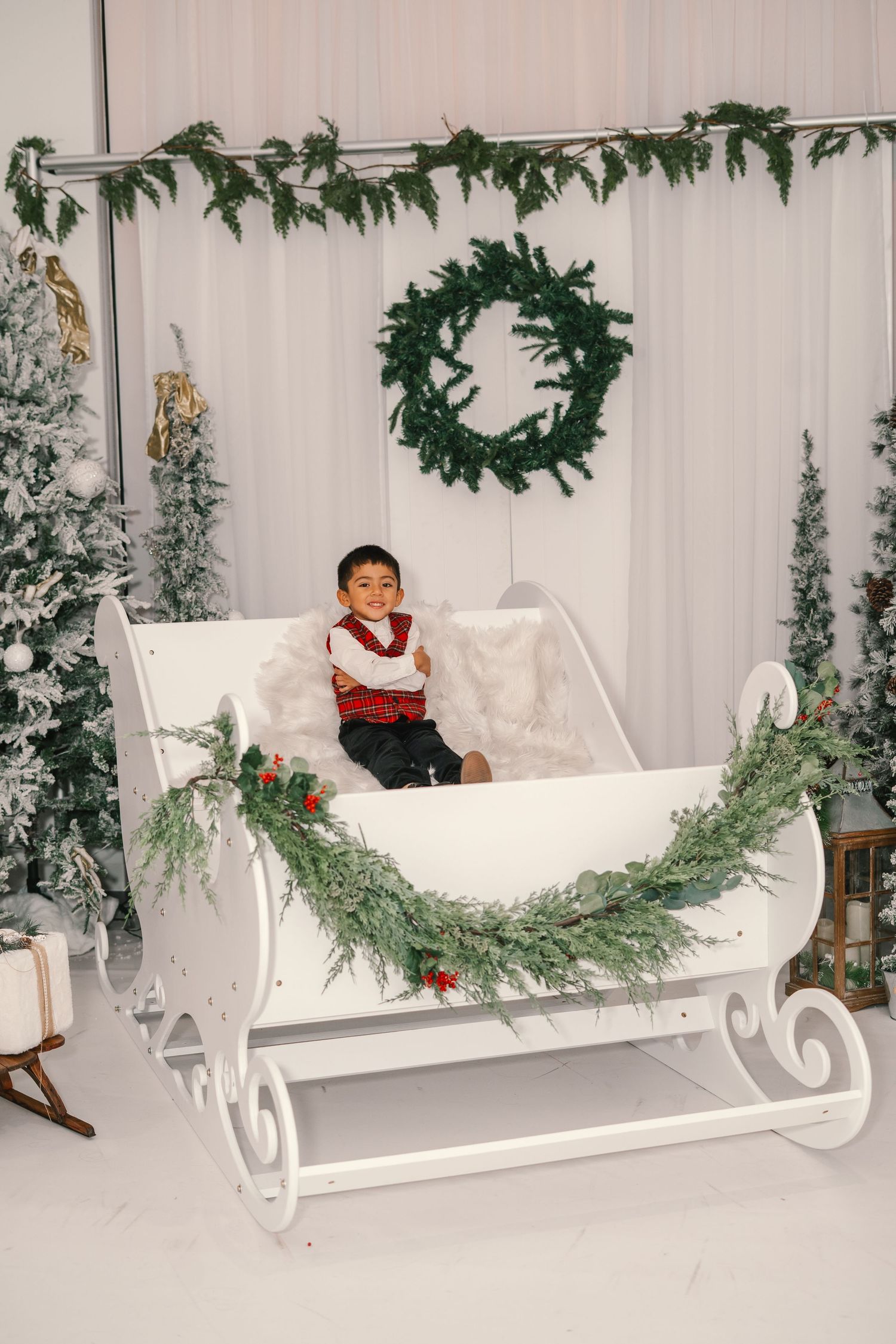 A young boy in a plaid vest sits alone in a white decorative sleigh surrounded by Christmas trees.
