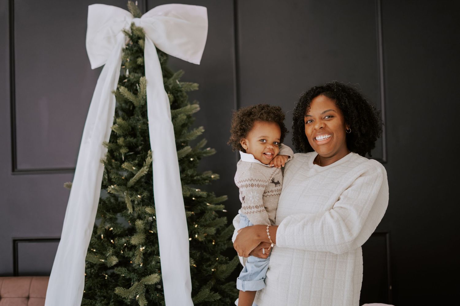 A smiling mother holds her baby boy in front of a decorated Christmas tree with a white bow.