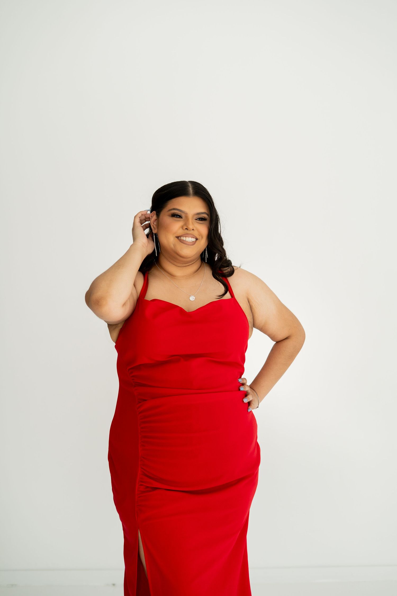 Woman in red spaghetti strap gown smiling with hand on hip against white background.