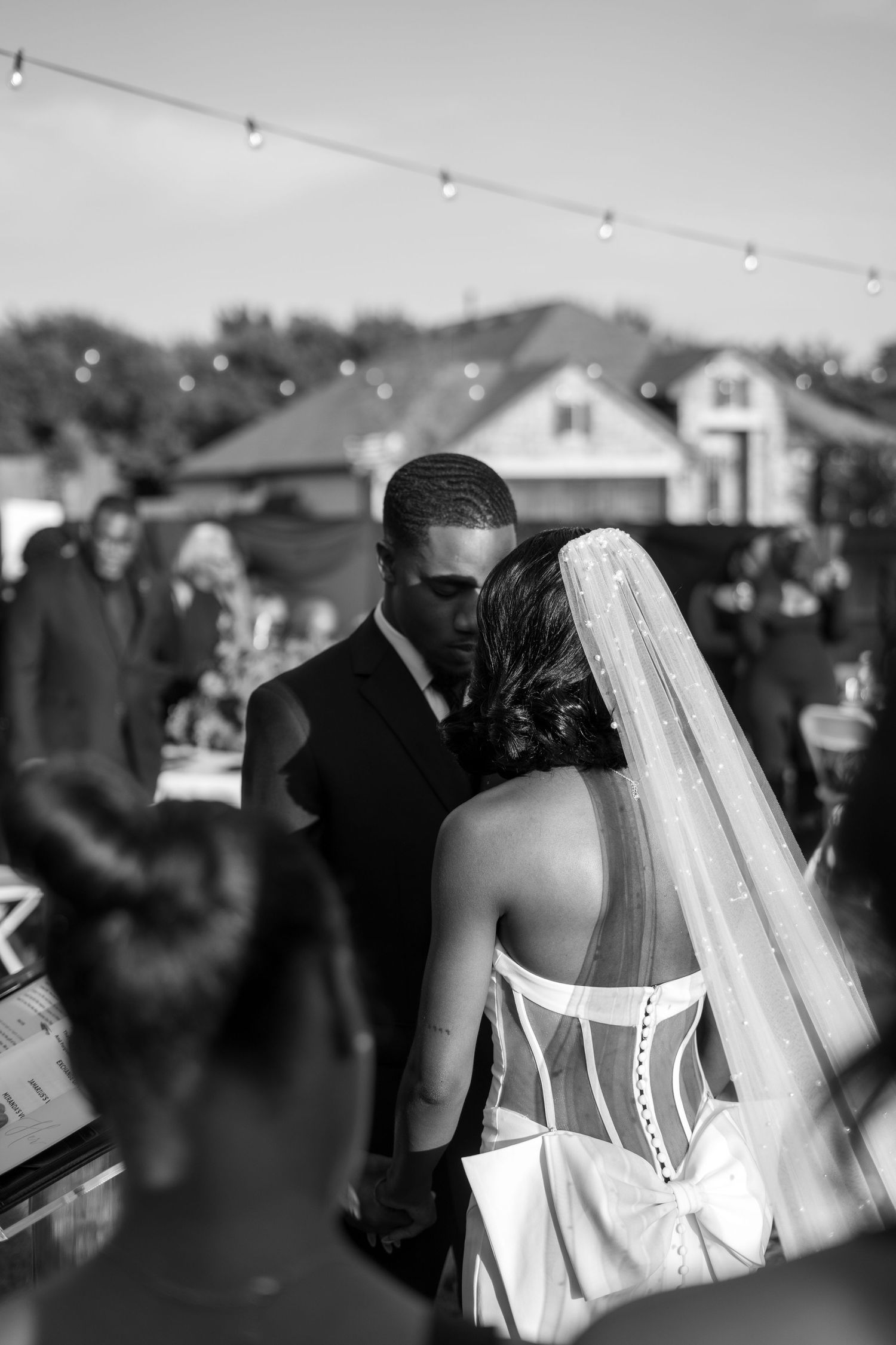 Black and white photo of bride and groom sharing an intimate moment during outdoor wedding ceremony with string lights.