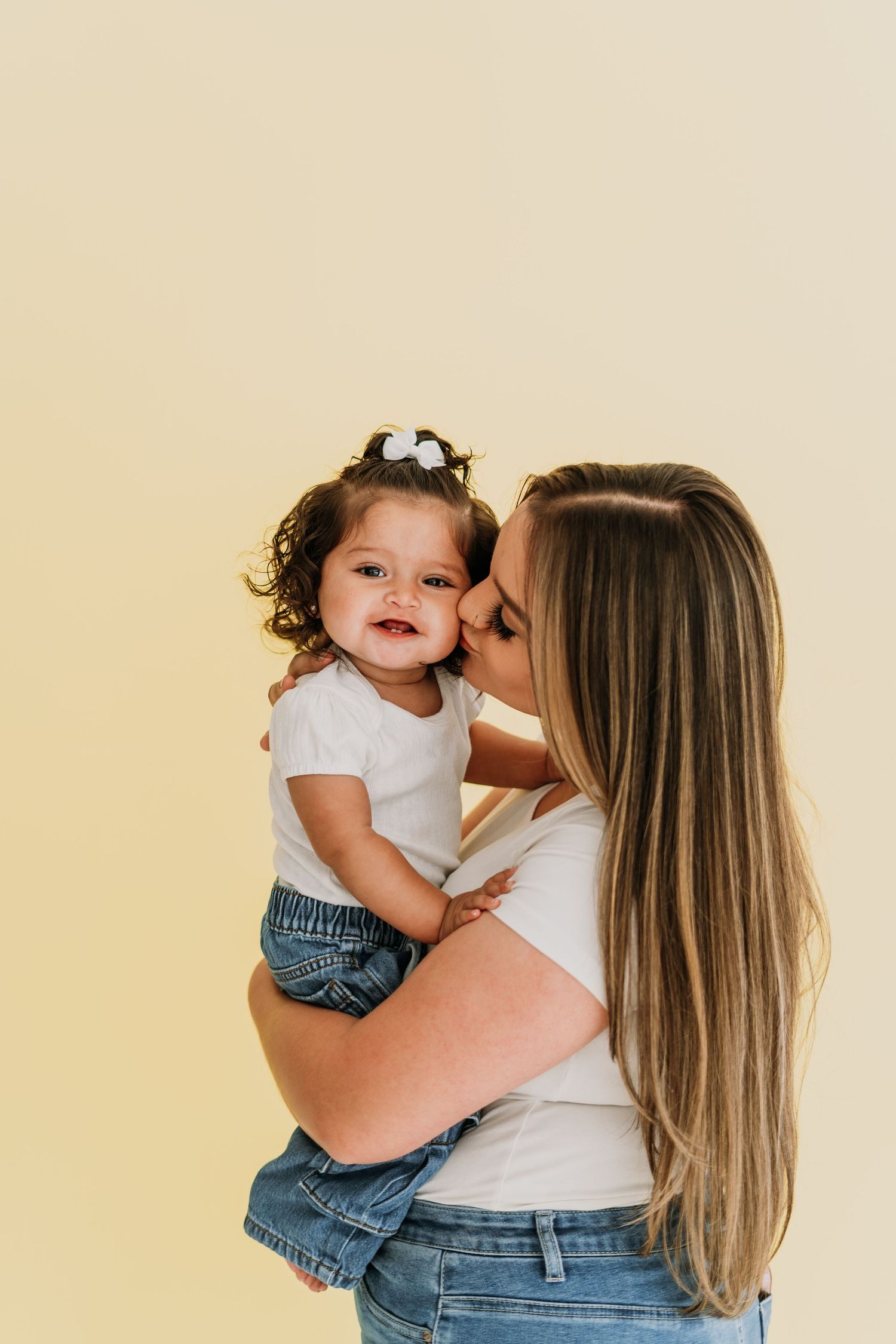 Mother kissing her curly-haired toddler daughter in white top and denim skirt against yellow wall.