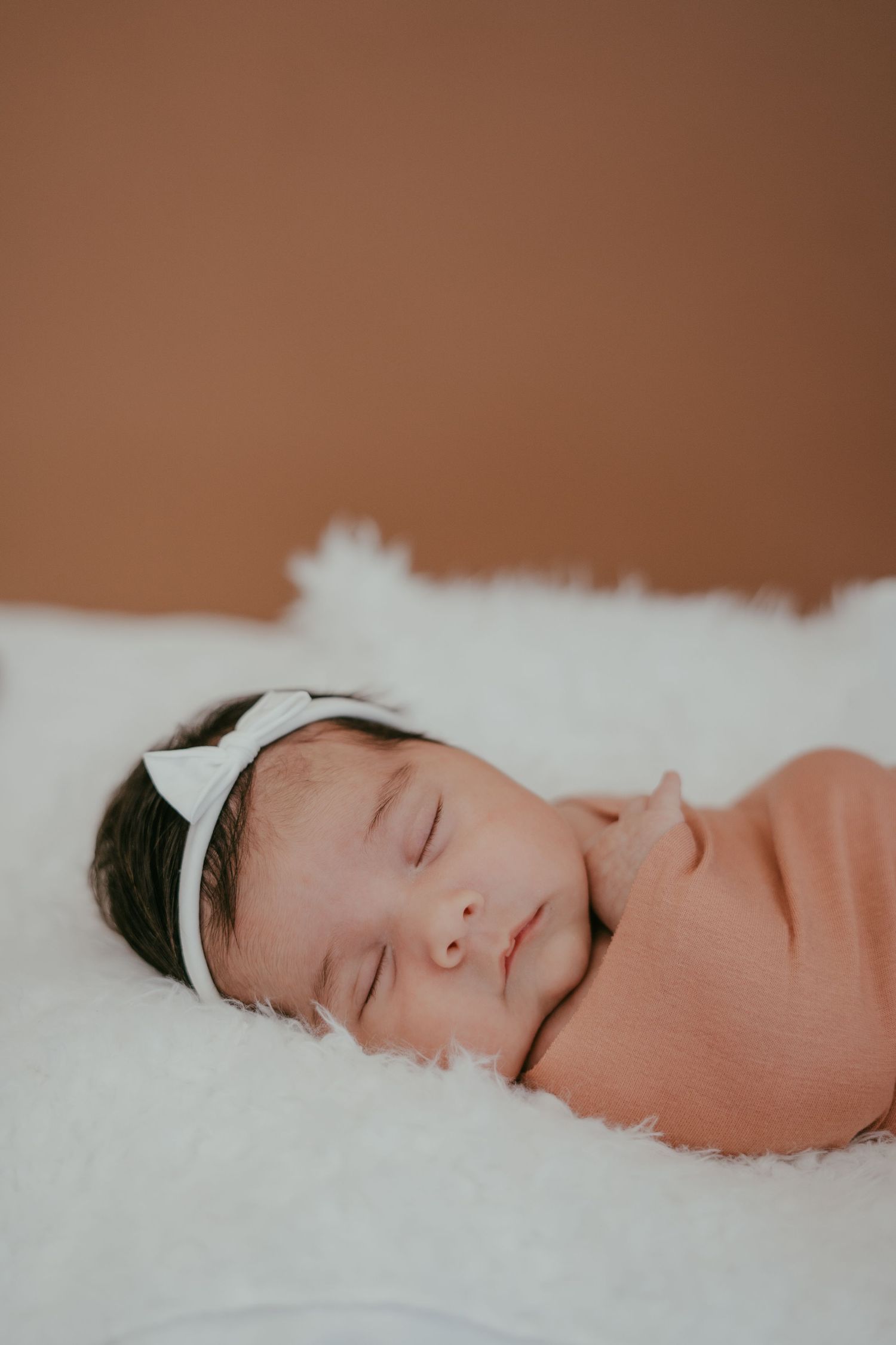 Serene newborn baby girl with white bow headband sleeps swaddled on a fluffy white blanket against brown background.