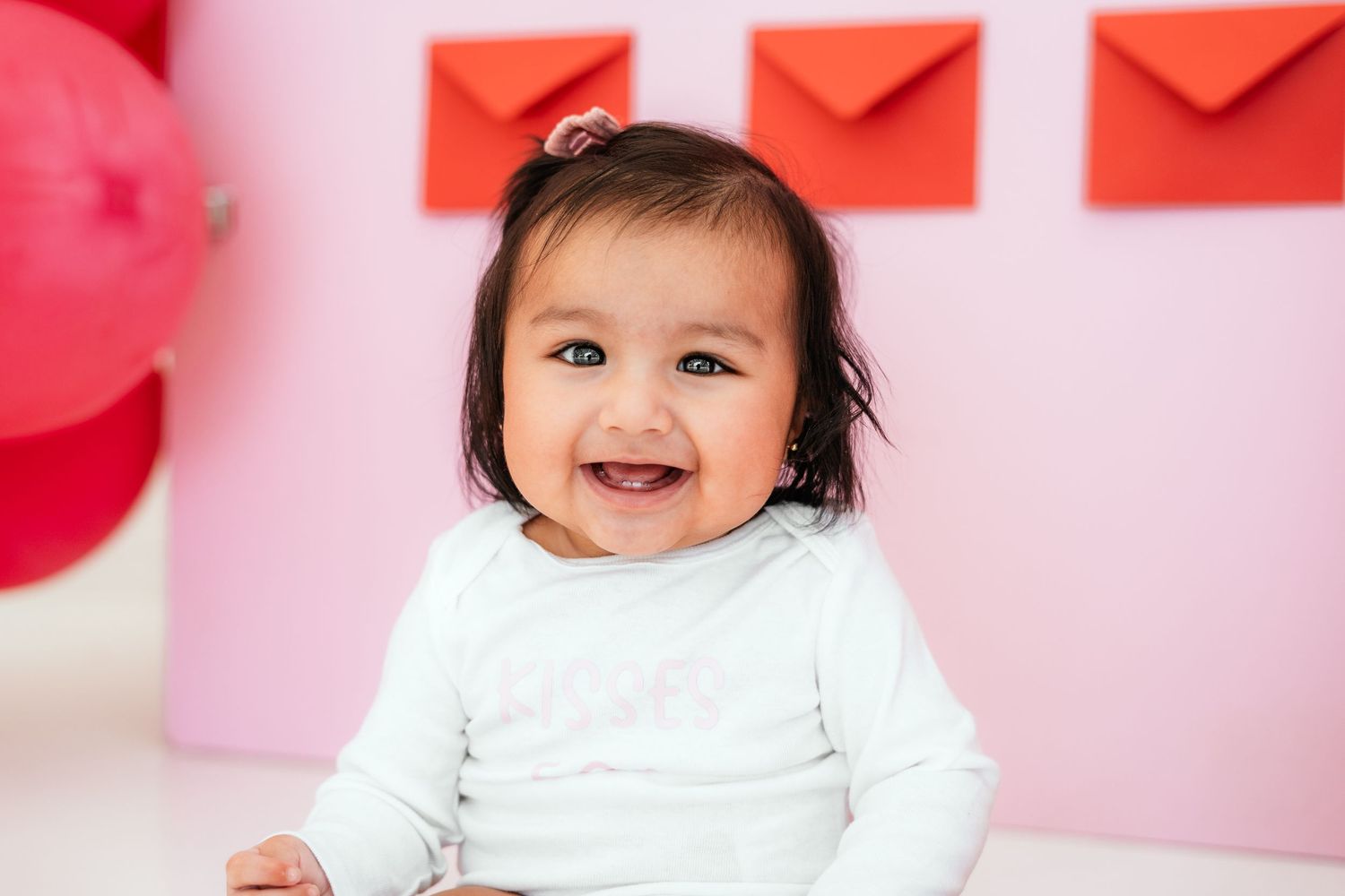 Smiling baby girl in white onesie poses before pink wall with red envelope decorations.