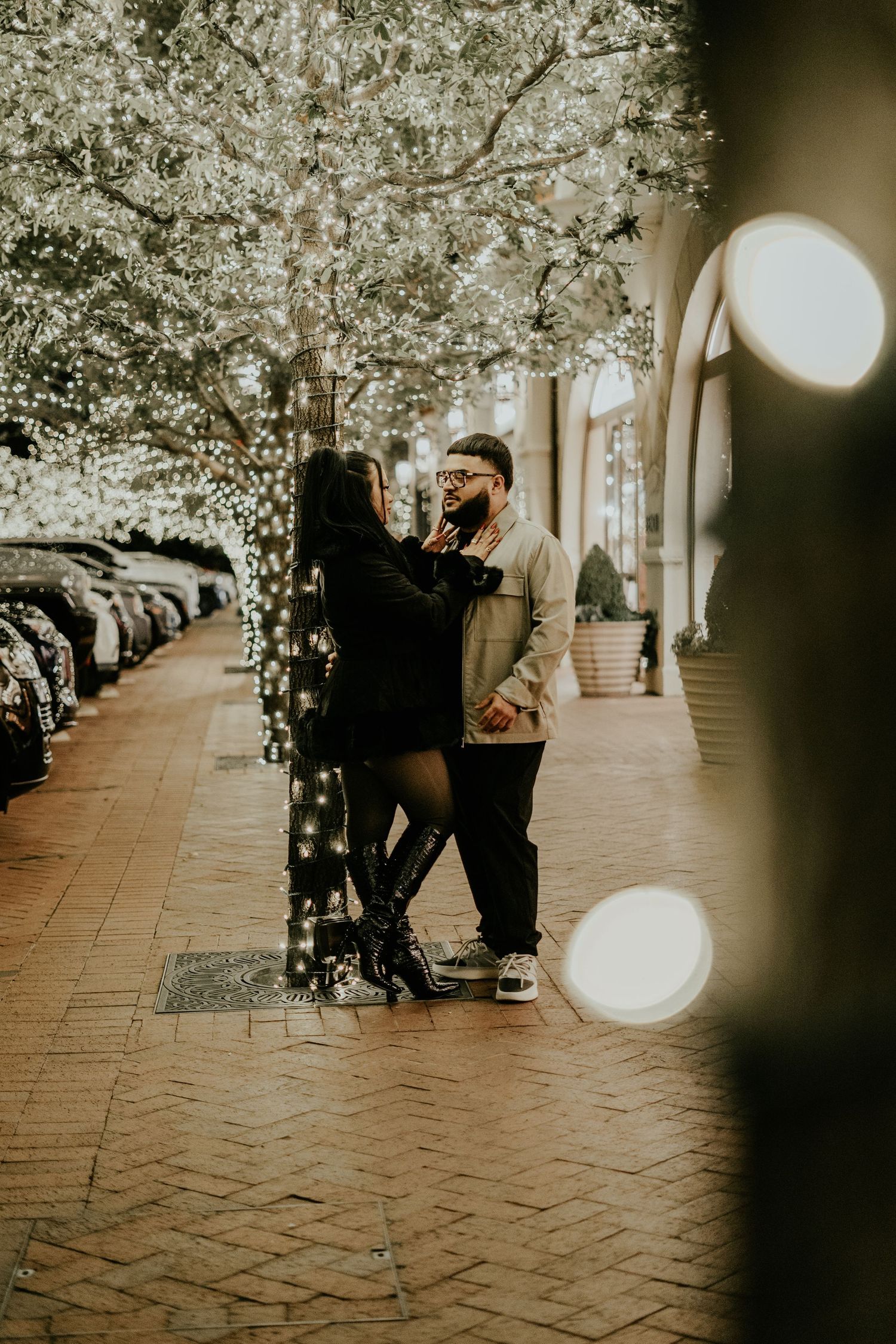 A couple stands close together under twinkling fairy lights on a romantic evening walkway.