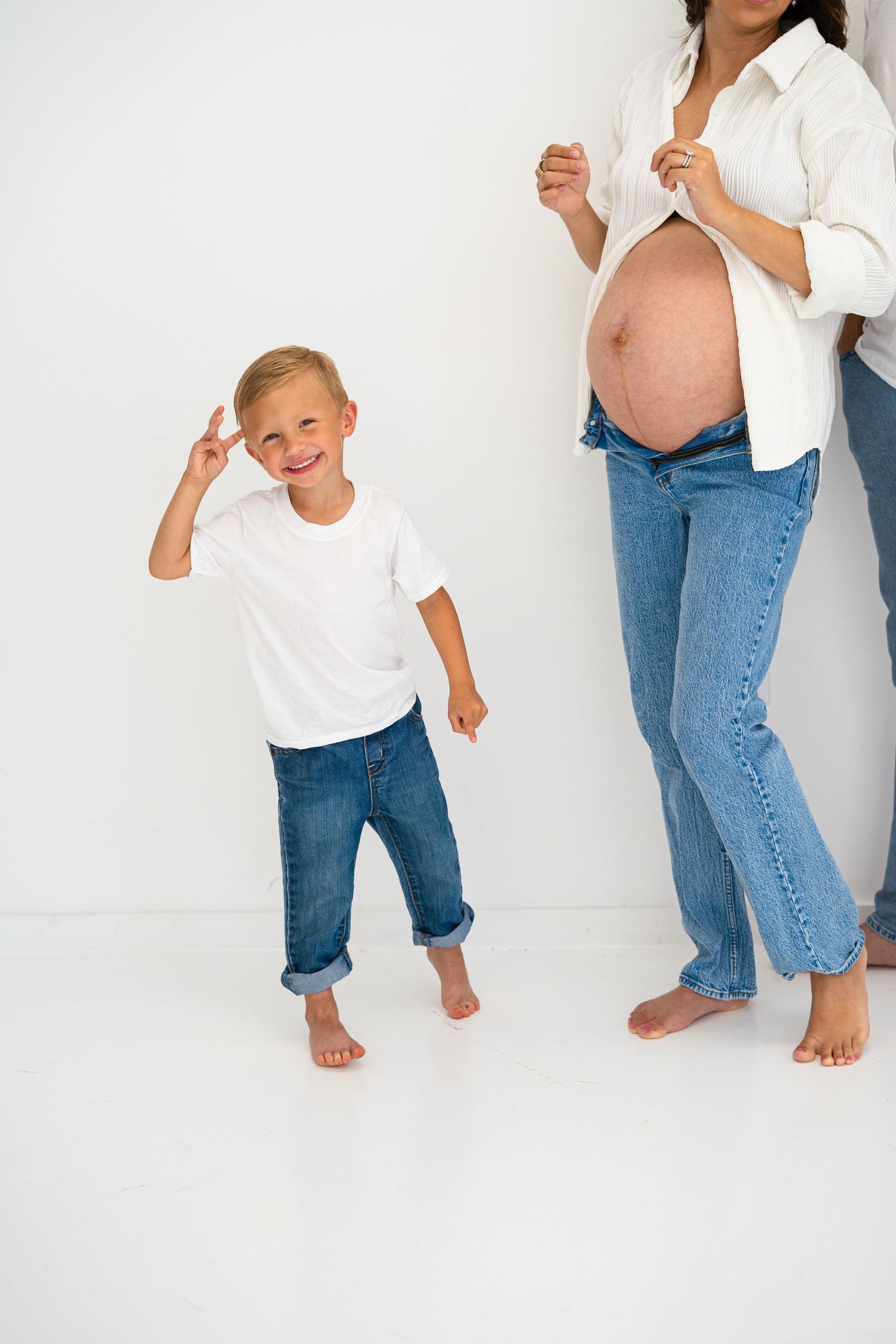A smiling blonde boy in a white shirt poses beside a pregnant woman showing her bare belly.