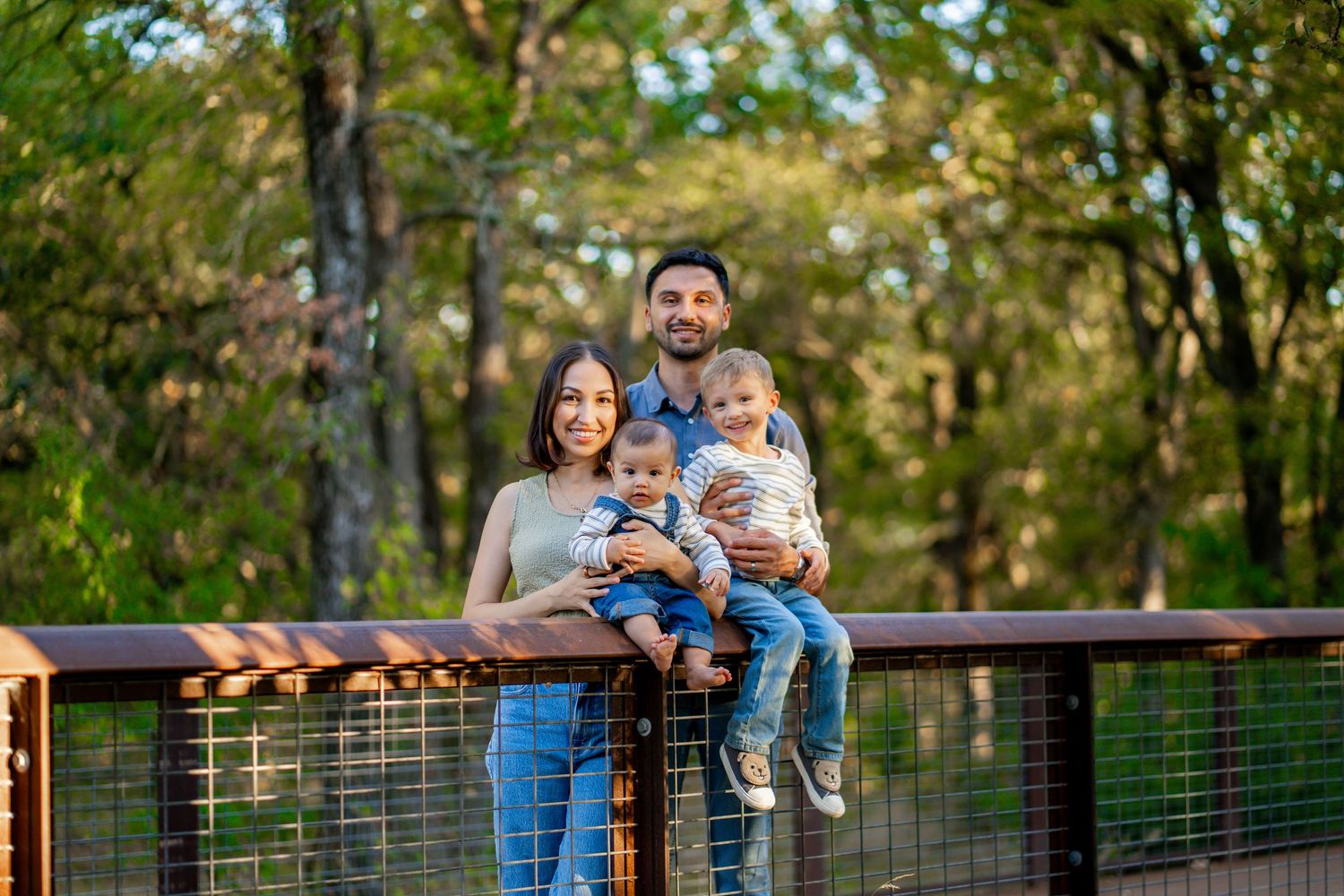 Smiling family of four posing on a wooden bridge railing surrounded by lush green forest.
