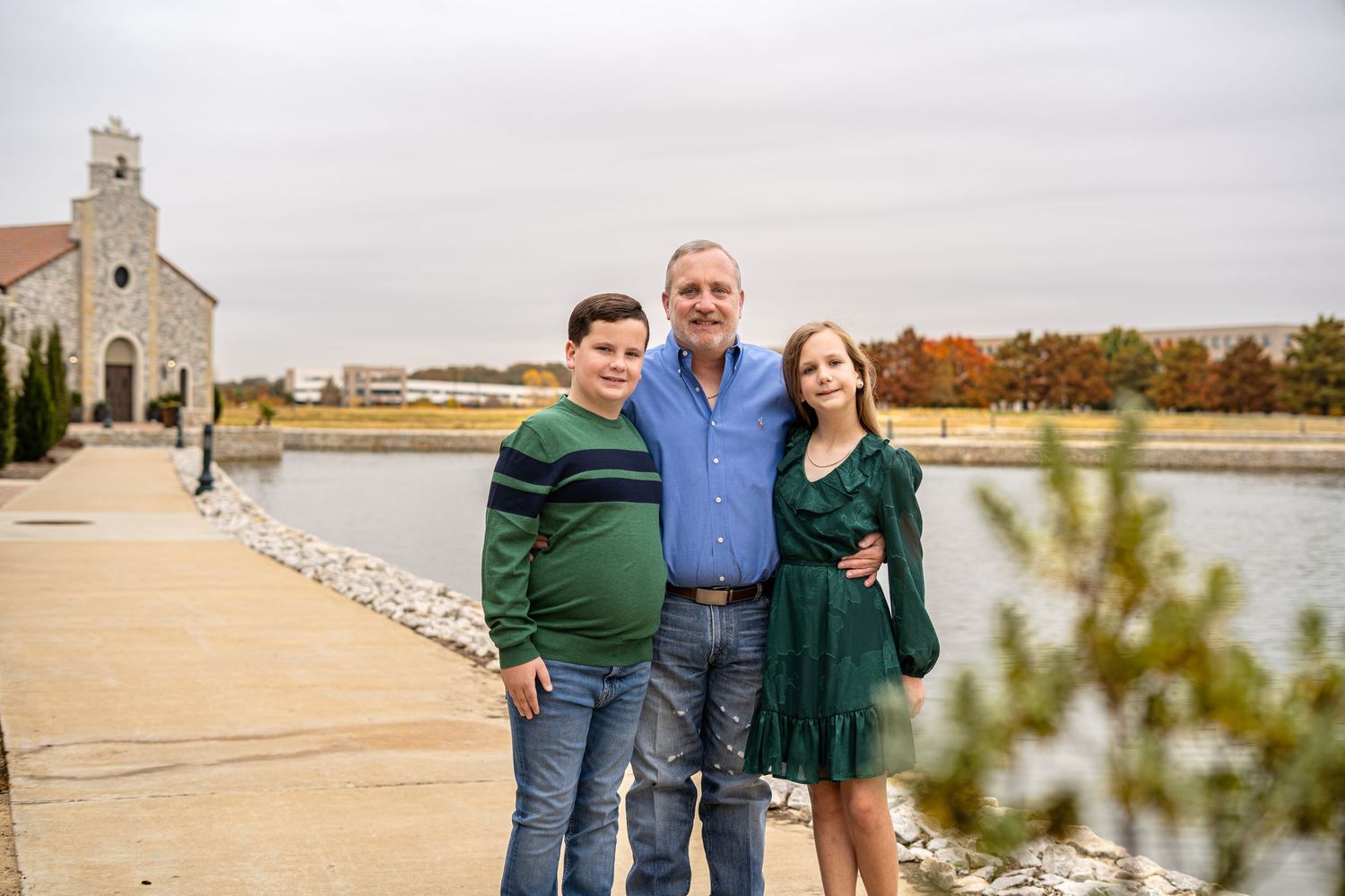 A man and two children smile together near a scenic waterfront with a stone church in the background.
