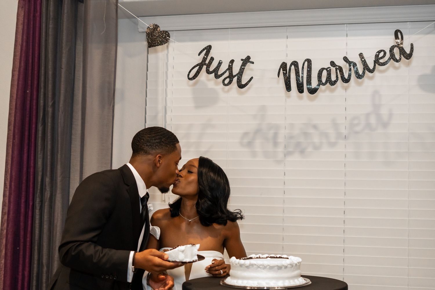 Newlywed couple sharing a romantic kiss beside their wedding cake under a Just Married banner.