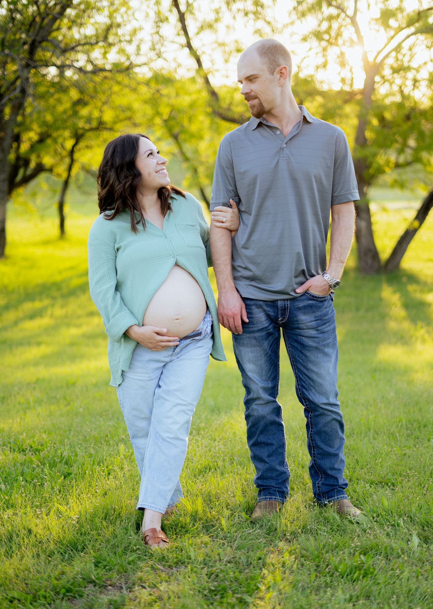 Pregnant couple smiling at each other while standing in lush green park during golden hour maternity photoshoot.