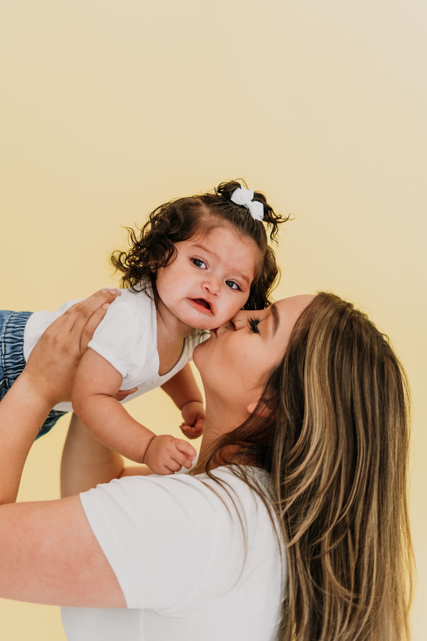 A mother lovingly kisses her curly-haired baby girl with a white bow, lifting her up against a soft yellow background.