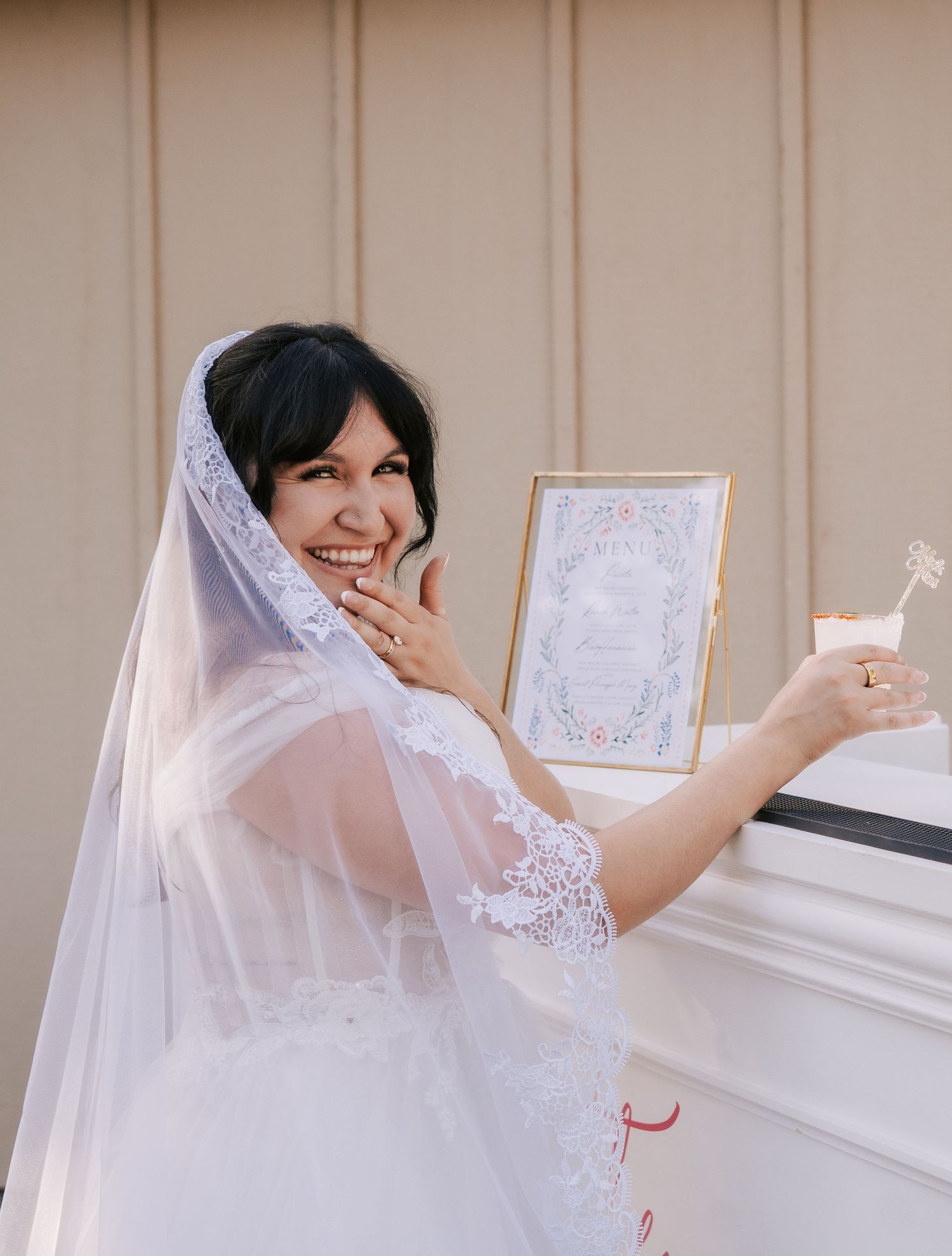 Smiling bride in white lace wedding dress and veil holding a framed sign and cocktail glass.