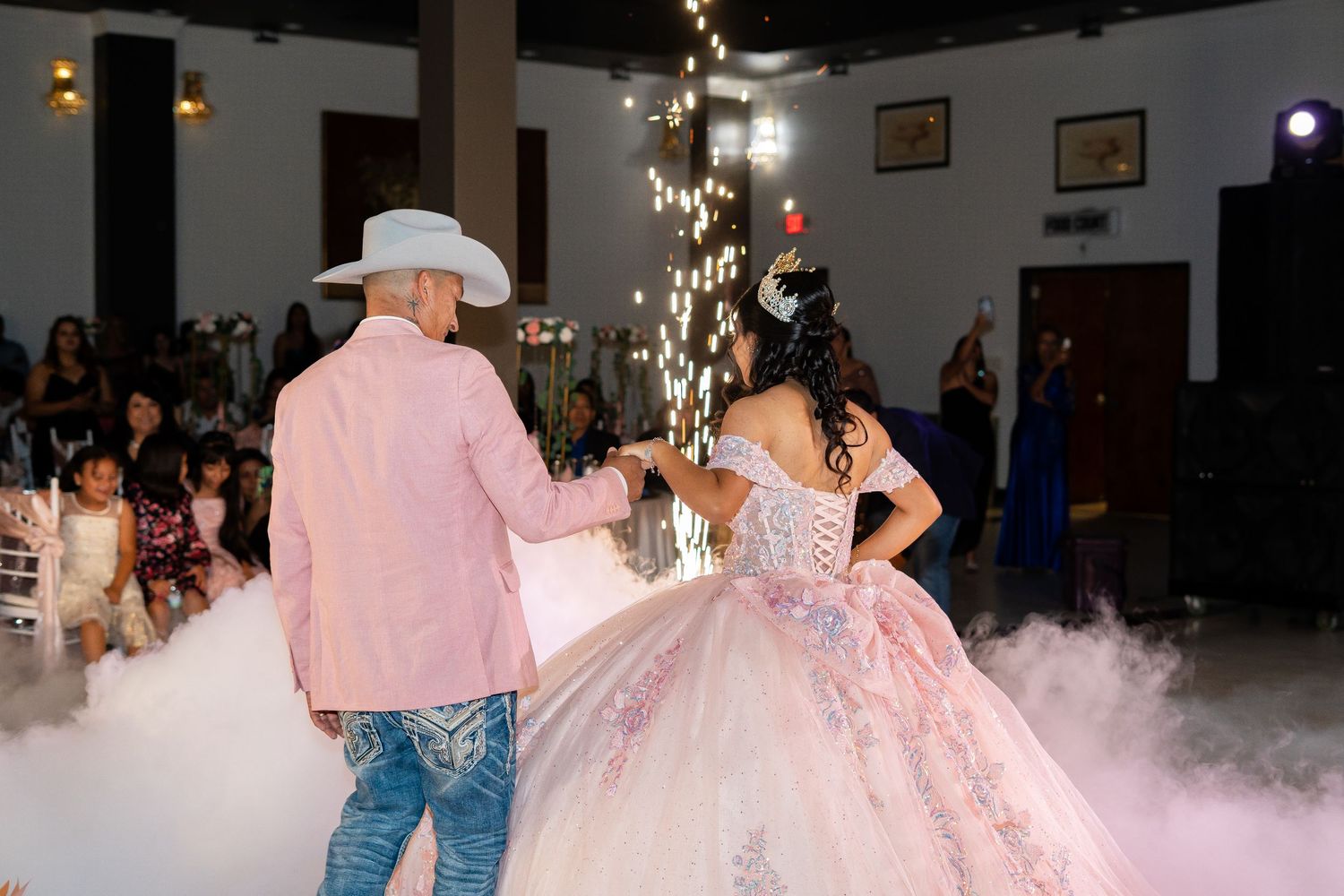 Quinceañera girl in pink ball gown dances with father in cowboy hat amid fog and sparkling lights.