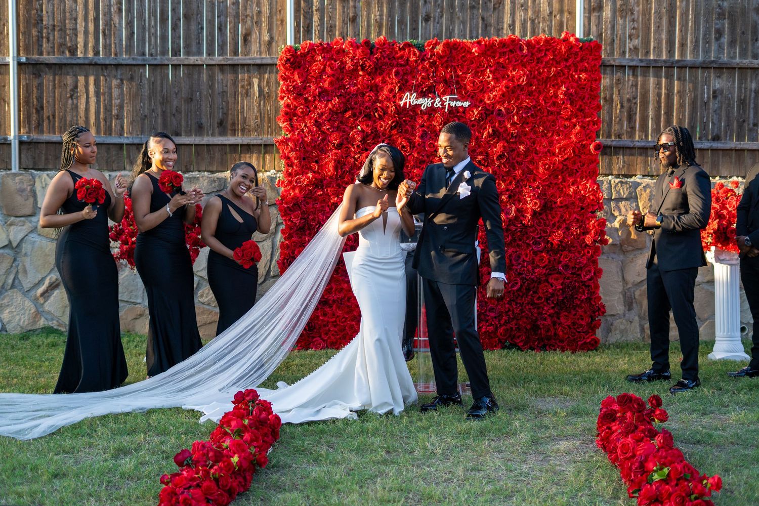 Newlyweds celebrate with wedding party in front of red rose wall after outdoor backyard ceremony.