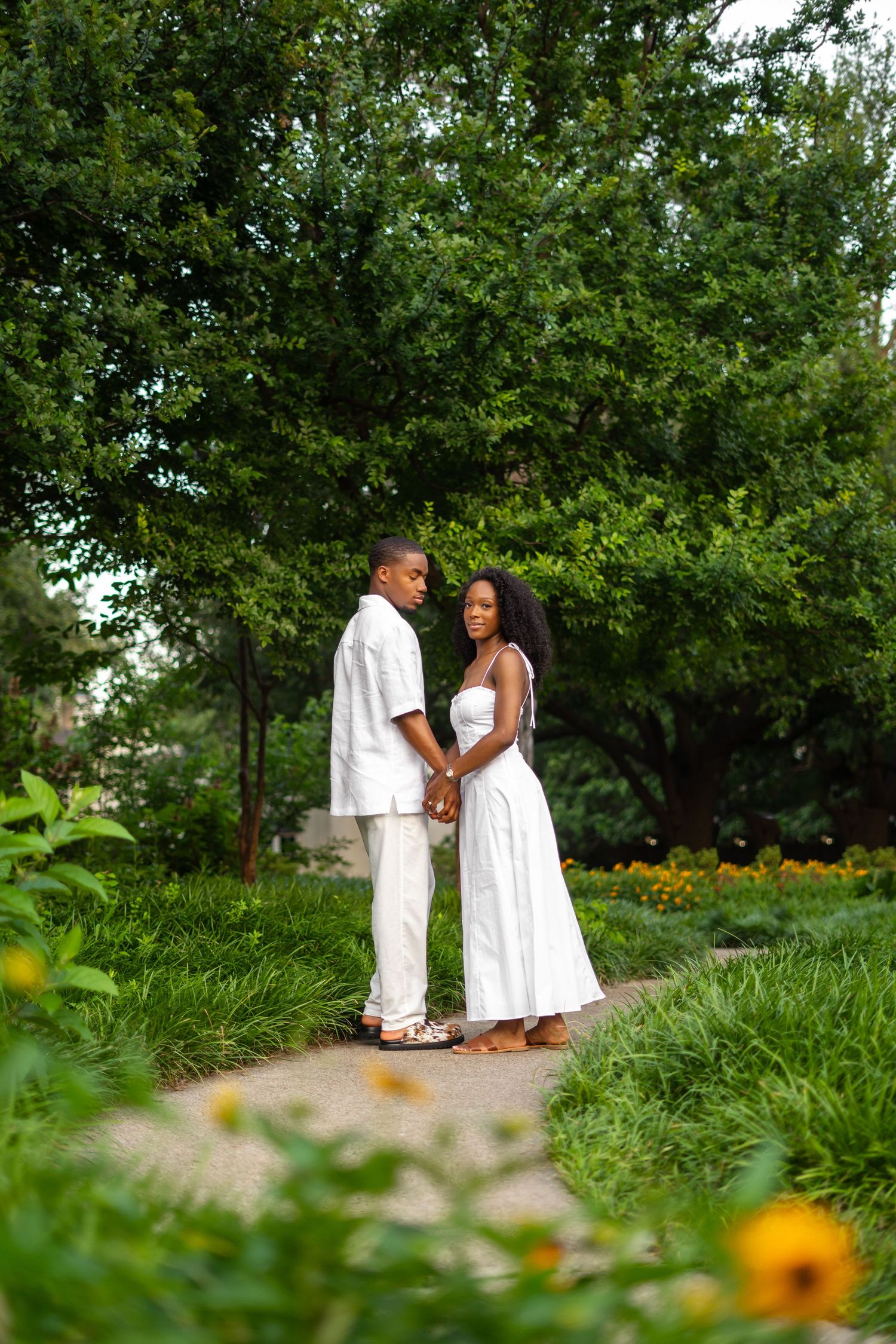 A romantic Black couple in white stands face-to-face holding hands on a garden path surrounded by greenery.