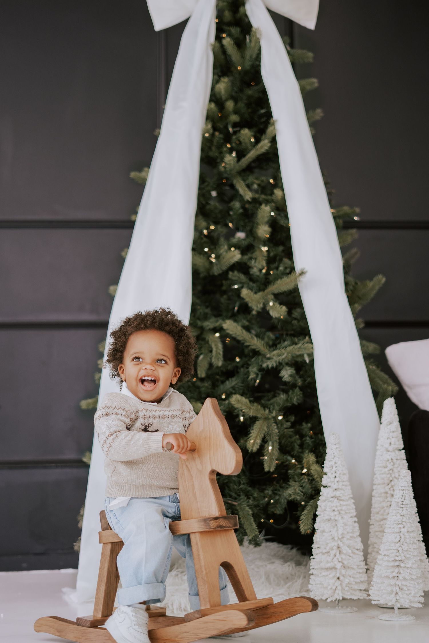 A laughing toddler rides a wooden rocking horse in front of a Christmas tree with a white bow.