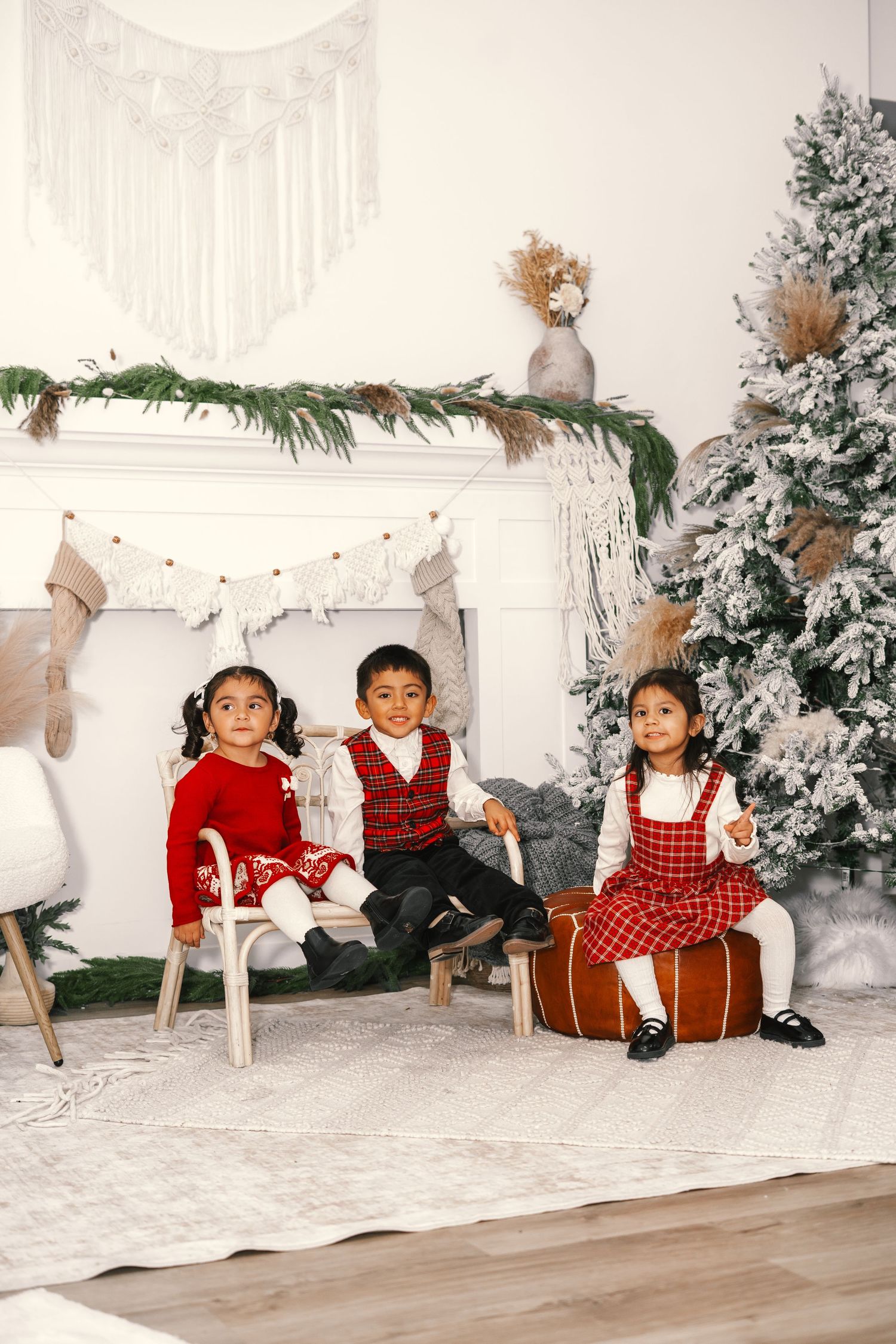Three young children in red holiday outfits pose together near a decorated Christmas tree and fireplace.