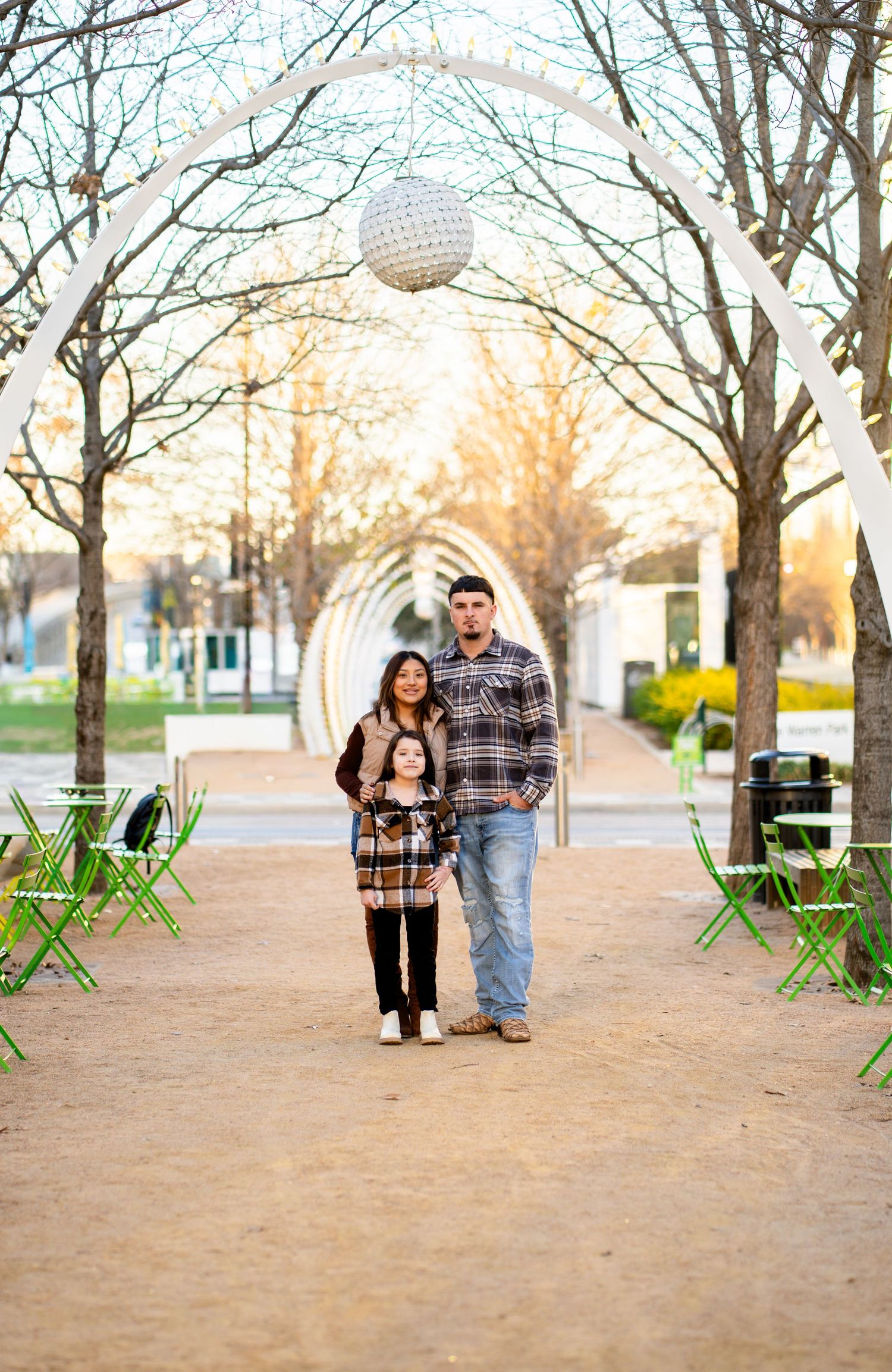 A family of three stands together on a scenic park pathway with arched decorations and warm sunset light.