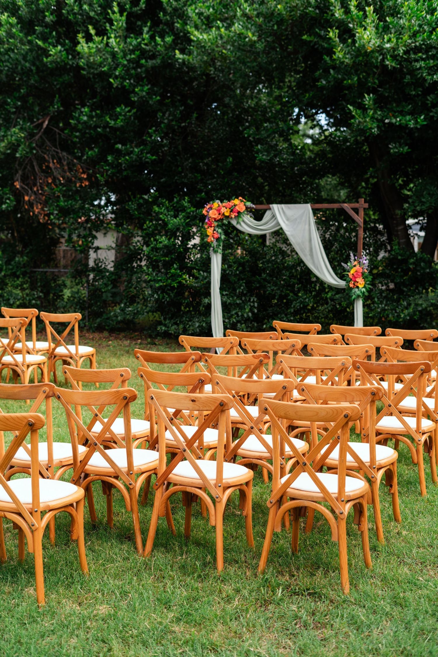 Outdoor wedding ceremony setup with wooden cross-back chairs on green grass and a floral arch with white draping.