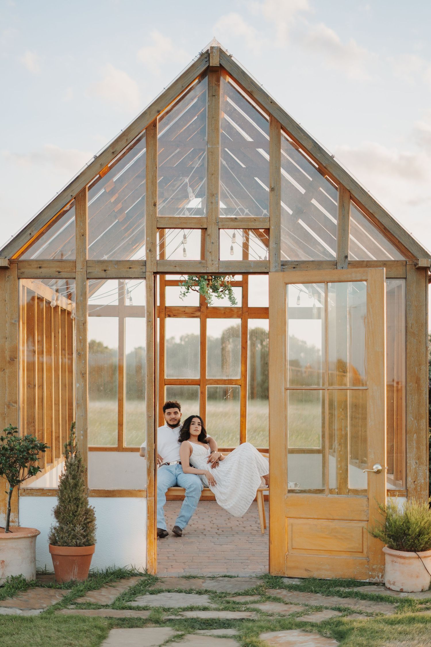 Engaged couple sits together inside a rustic wooden greenhouse at golden hour on a sunny day.