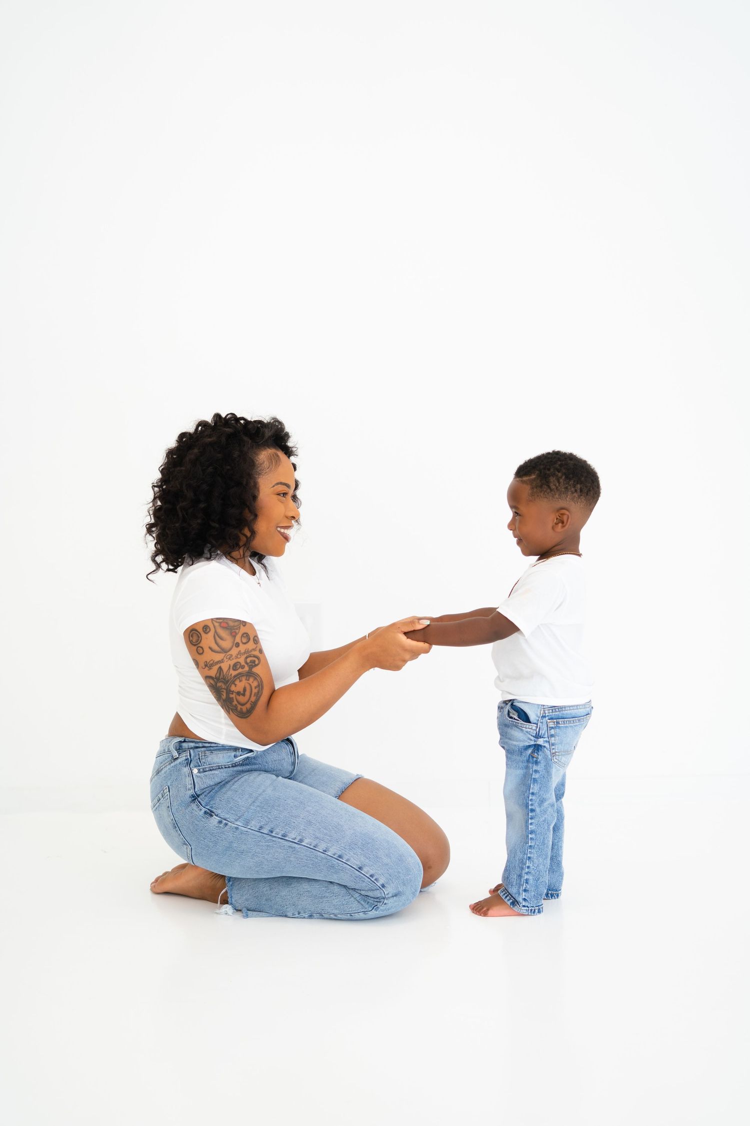 Tattooed Black mother kneeling and holding hands with her young son in white studio setting.
