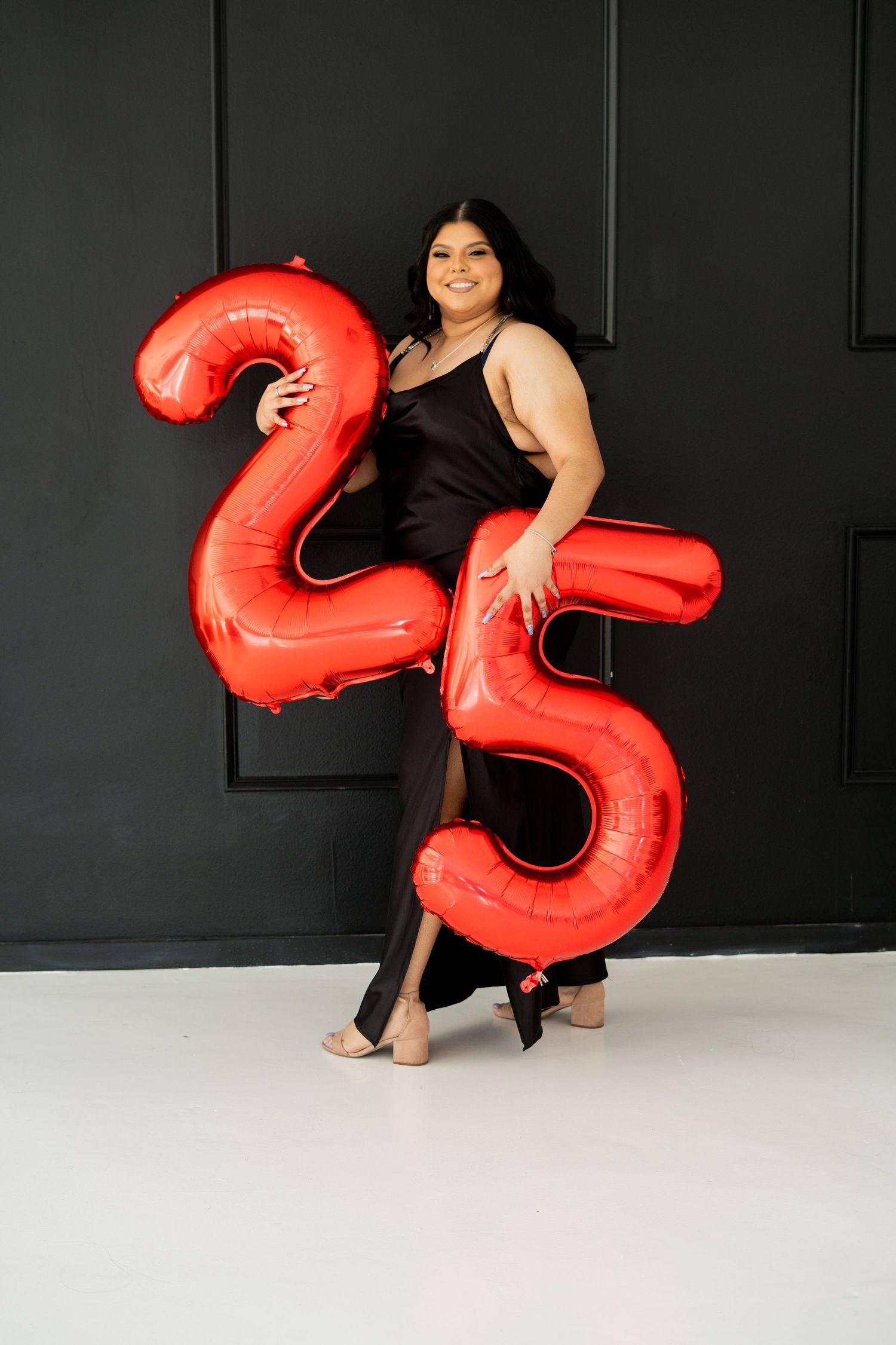 A woman in a black dress holds large red number 25 balloons celebrating her birthday.