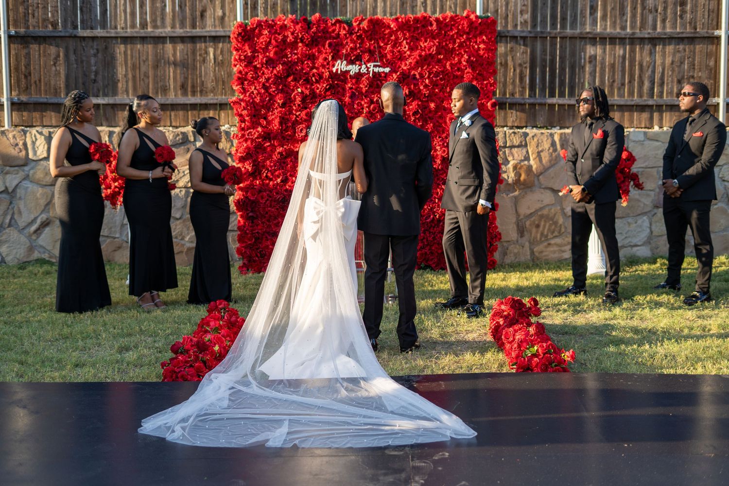 Bride and groom stand before red rose flower wall altar surrounded by wedding party at outdoor ceremony.