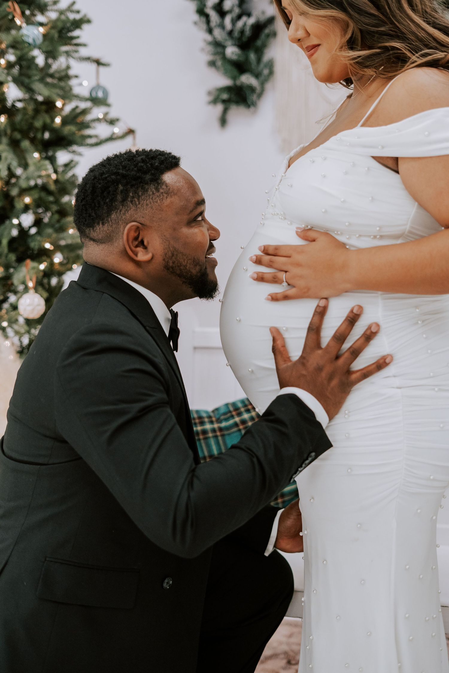 Groom in black tuxedo kneeling to kiss pregnant bride's baby bump near decorated Christmas tree.